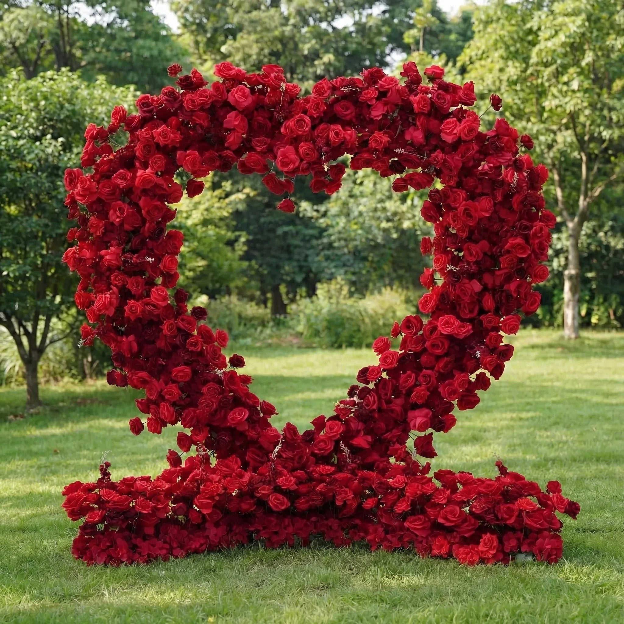This is a striking heart-shaped artificial floral arch from **Valar Flowers**, entirely covered in lush, vibrant red roses. Set on a lush green lawn with dense trees in the background, it creates a bold, romantic, and eye-catching aesthetic ideal for marriage proposals, wedding ceremonies, or special photo backdrops.
