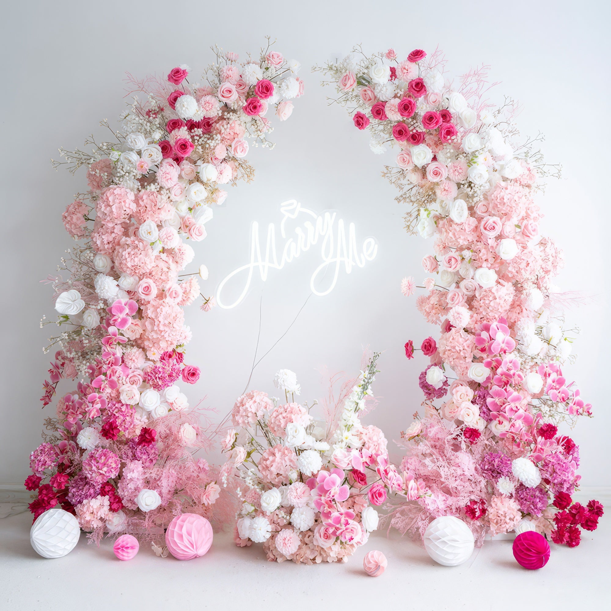 This romantic floral arch, adorned with a lush gradient of blush pink, deep rose, and white blooms, is paired with a glowing "Marry Me" neon sign at its center. The arrangement, featuring roses, hydrangeas, and delicate filler flowers, creates a dreamy, three-dimensional texture. Set against a minimalist white backdrop and accented with pink and white paper lanterns, it exudes timeless romance, making it the perfect proposal or engagement photoshoot focal point.