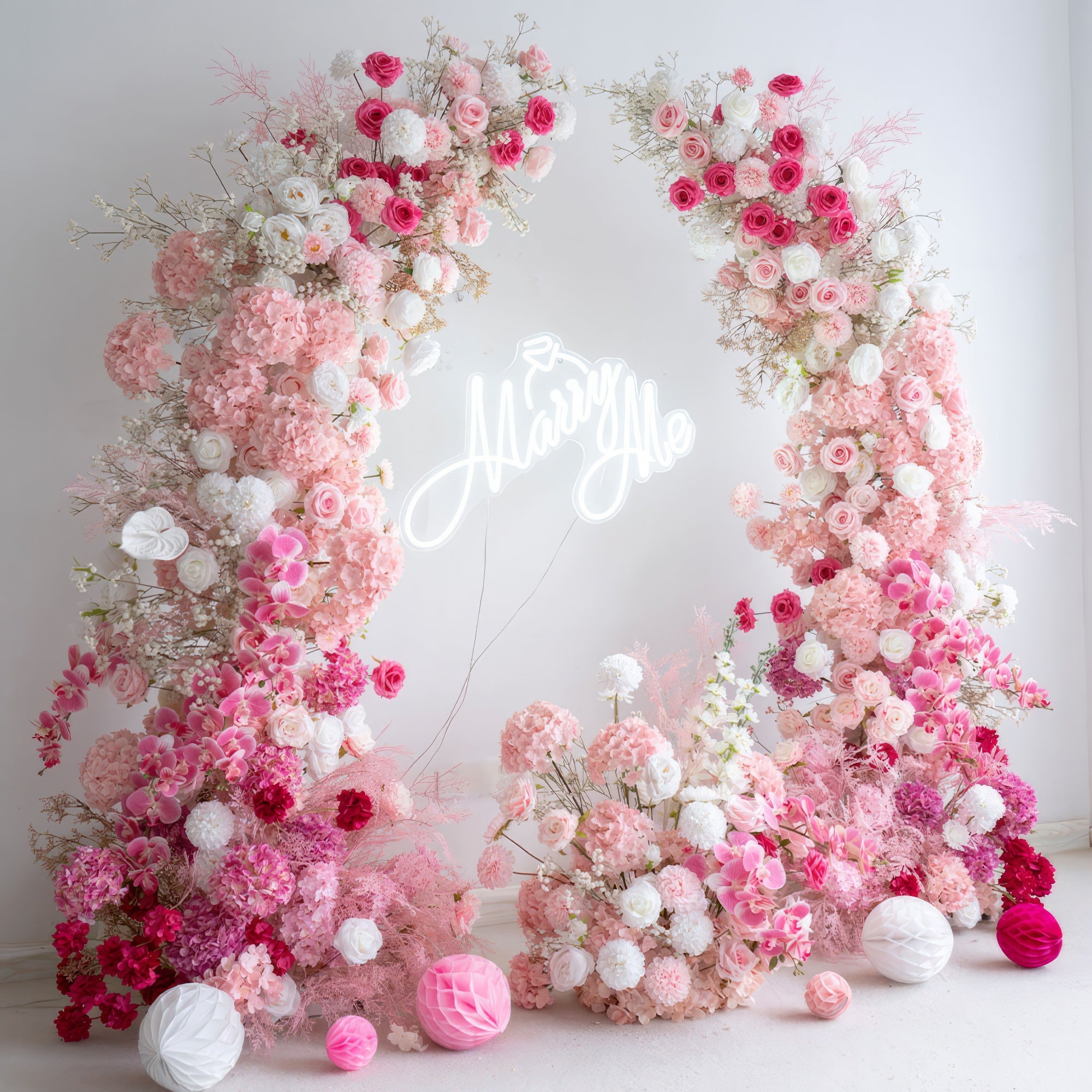 This romantic proposal-ready floral arch is adorned with a lush gradient of blush pink, deep rose, and white blooms, including roses, hydrangeas, and delicate filler flowers. At its center, a glowing "Marry Me" neon sign takes the spotlight. The arrangement creates a dreamy, three-dimensional texture, and is accented with pink and white paper lanterns. Set against a minimalist white backdrop, it exudes timeless romance, making it the perfect focal point for a heartfelt proposal or engagement photoshoot.