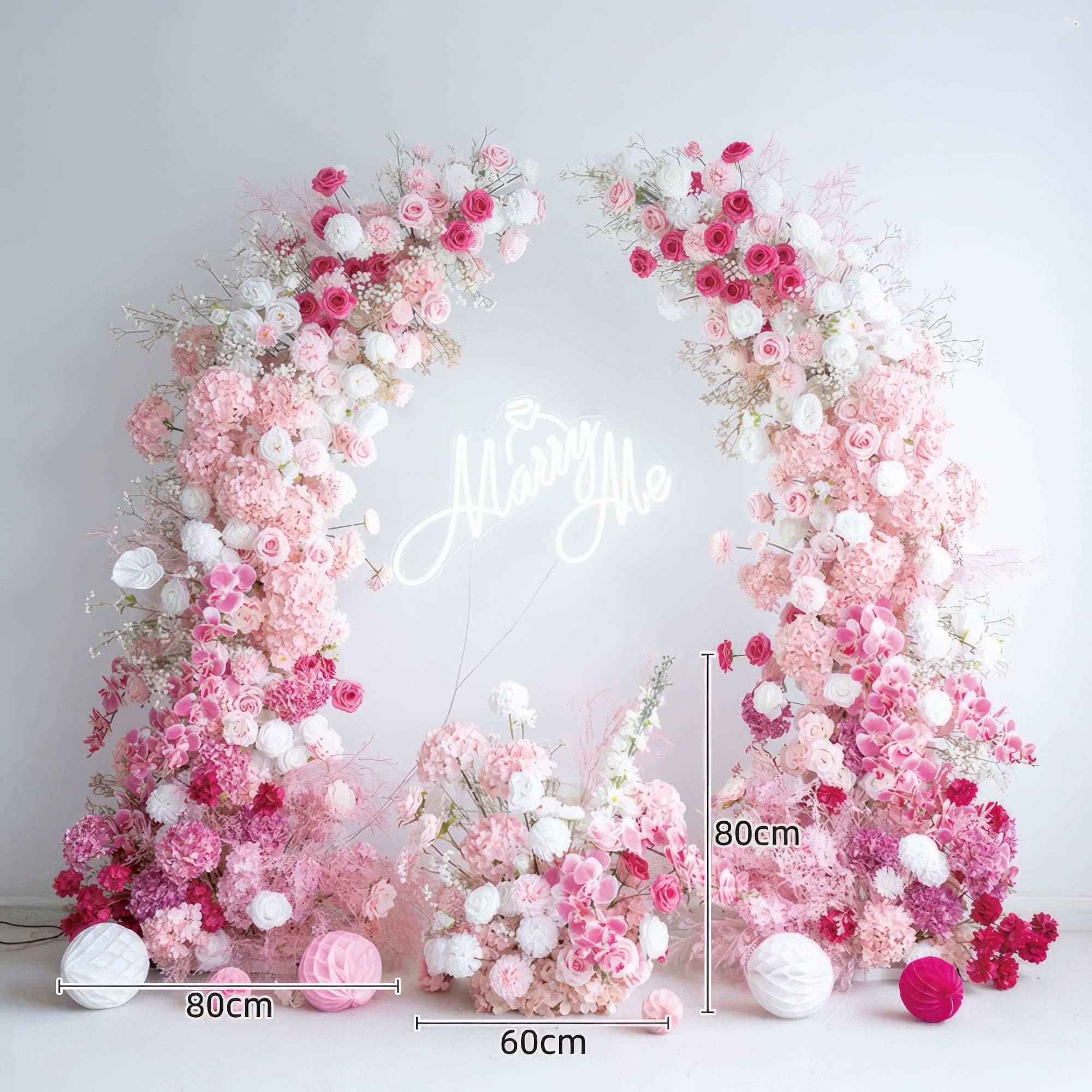 This romantic, proposal-ready floral arch is adorned with a lush gradient of blush pink, deep rose, and white blooms, including roses, hydrangeas, and delicate filler flowers. At its center, a glowing "Marry Me" neon sign takes the spotlight. The arrangement creates a dreamy, three-dimensional texture, and is accented with pink and white paper lanterns. Set against a minimalist white backdrop, it exudes timeless romance, making it the perfect focal point for a heartfelt proposal or engagement photoshoot.