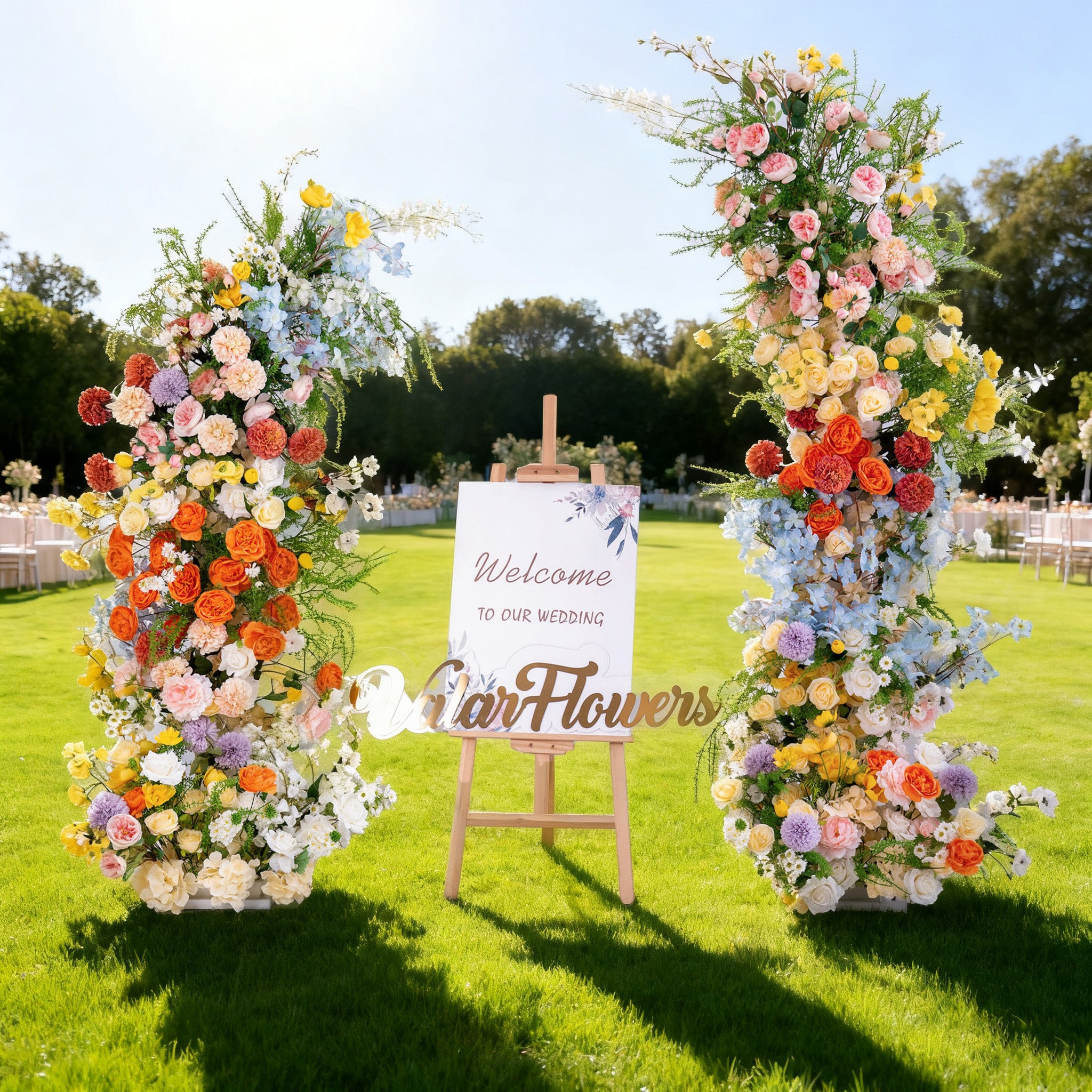 This image captures a charming outdoor wedding scene. Two magnificent floral arrangements in vibrant hues of orange, yellow, pink, blue, and purple flank a wooden easel. The easel holds a sign that reads "Welcome TO OUR WEDDING" with "Valar Flowers" below. Set on a lush green lawn under a bright, sunny sky with trees in the background, the scene exudes a joyful and romantic atmosphere, perfect for a wedding celebration.