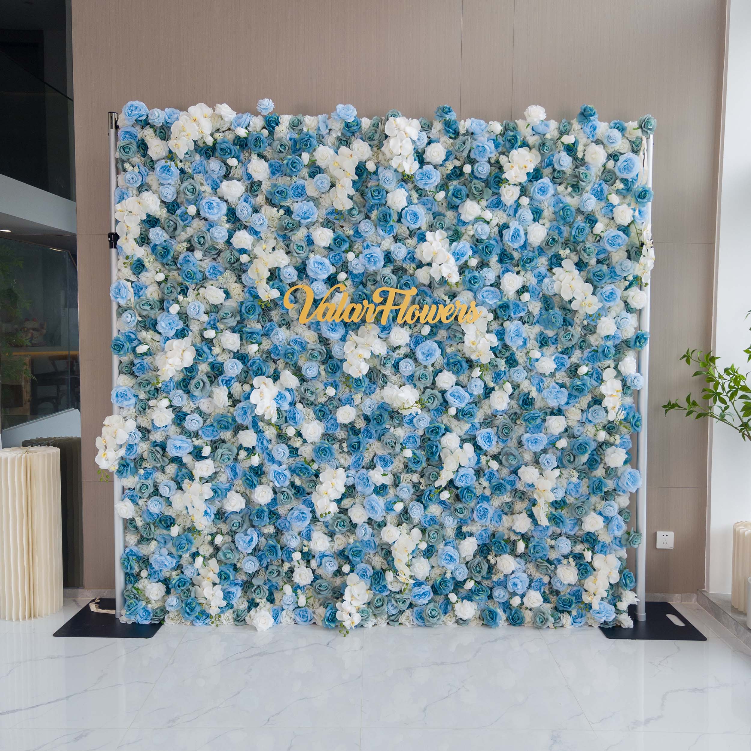 This photo showcases a large, eye - catching floral backdrop densely covered with blue and white roses and white orchids. The flowers create a lush, textured appearance. Prominently displayed in the center is the golden text "ValarFlowers," adding a touch of branding. Set against a neutral wall and a polished marble floor, the backdrop exudes elegance, making it perfect for upscale events like weddings or corporate galas.