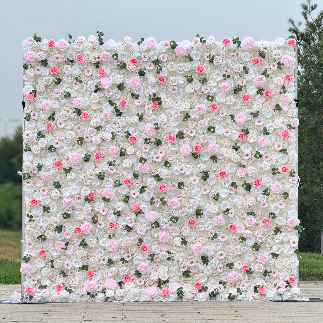 This photo showcases a stunning floral wall, likely by ValarFlowers, featuring artificial roses in soft pink and white hues, interspersed with greenery. The dense arrangement creates a romantic and elegant ambiance, perfect for weddings or other formal events. The realistic appearance of the faux flowers adds a touch of timeless beauty, making it an ideal backdrop for capturing memorable moments with a dreamy, celebratory atmosphere.