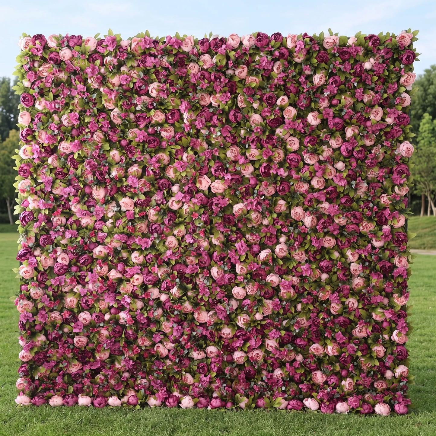 Full-size artificial flower wall boasting dense layered blooms in blush pink, lavender, magenta and deep plum roses mixed with green foliage, displayed on a sunlit lawn. This maintenance-free reusable floral backdrop creates rich romantic texture, ideal for wedding photo booths, bridal events, boutique retail decor and outdoor garden parties, delivering vibrant Instagram-worthy pink-purple tones for all celebration styling.