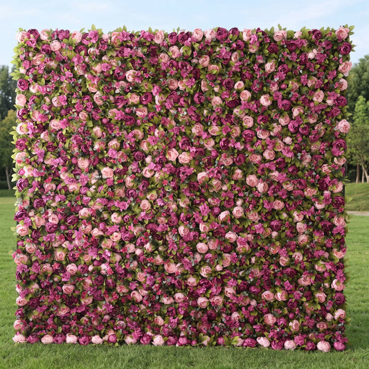 Full-size artificial flower wall boasting dense layered blooms in blush pink, lavender, magenta and deep plum roses mixed with green foliage, displayed on a sunlit lawn. This maintenance-free reusable floral backdrop creates rich romantic texture, ideal for wedding photo booths, bridal events, boutique retail decor and outdoor garden parties, delivering vibrant Instagram-worthy pink-purple tones for all celebration styling.