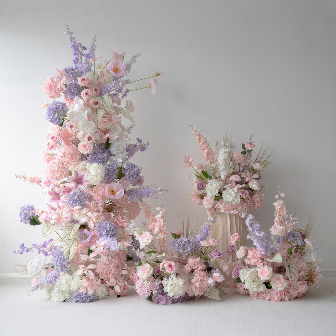 This set of floral decorations features a soft and dreamy color scheme with shades of pink, purple, and white. There are various artificial flowers such as roses, hydrangeas, and lilies, along with some delicate foliage. The large vertical arrangement and smaller pieces on the ground create a layered and romantic look, ideal for a wedding ceremony, bridal shower, or other elegant events.