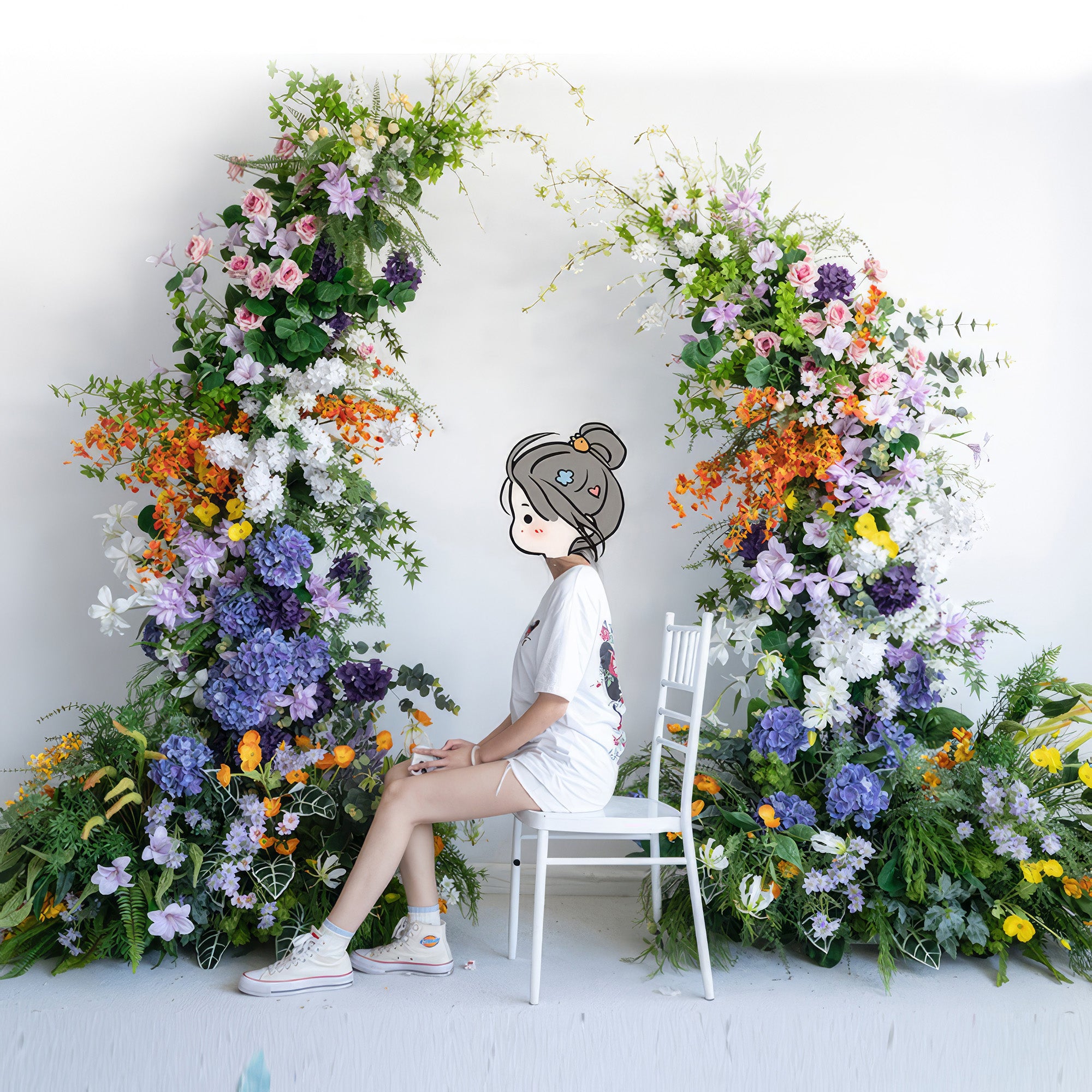 This image showcases a vivid artificial floral arch with a diverse color scheme of purple, pink, orange, white, and yellow. It includes different flower types like hydrangeas and roses. A person sits on a white chair in front, creating a lively and charming scene. 