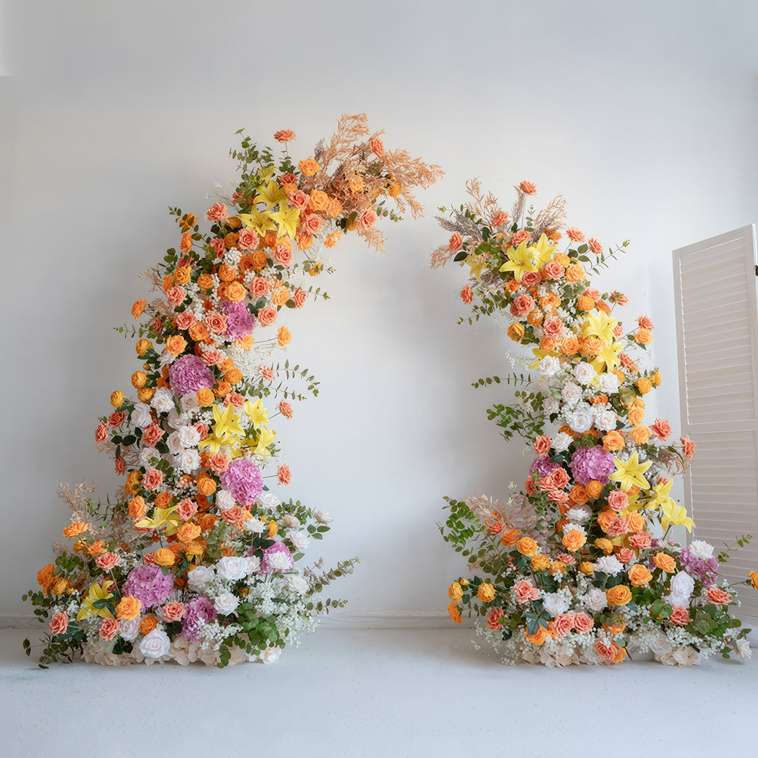 This floral arch is a vibrant display of colors, featuring a mix of orange, yellow, pink, and white flowers. There are roses, hydrangeas, lilies, and other blooms, complemented by green foliage and some dried elements. It creates a lively and eye - catching look, perfect for adding a festive touch to weddings, parties, or other special events.