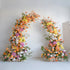 This floral arch is a vibrant display of colors, featuring a mix of orange, yellow, pink, and white flowers. There are roses, hydrangeas, lilies, and other blooms, complemented by green foliage and some dried elements. It creates a lively and eye - catching look, perfect for adding a festive touch to weddings, parties, or other special events. 