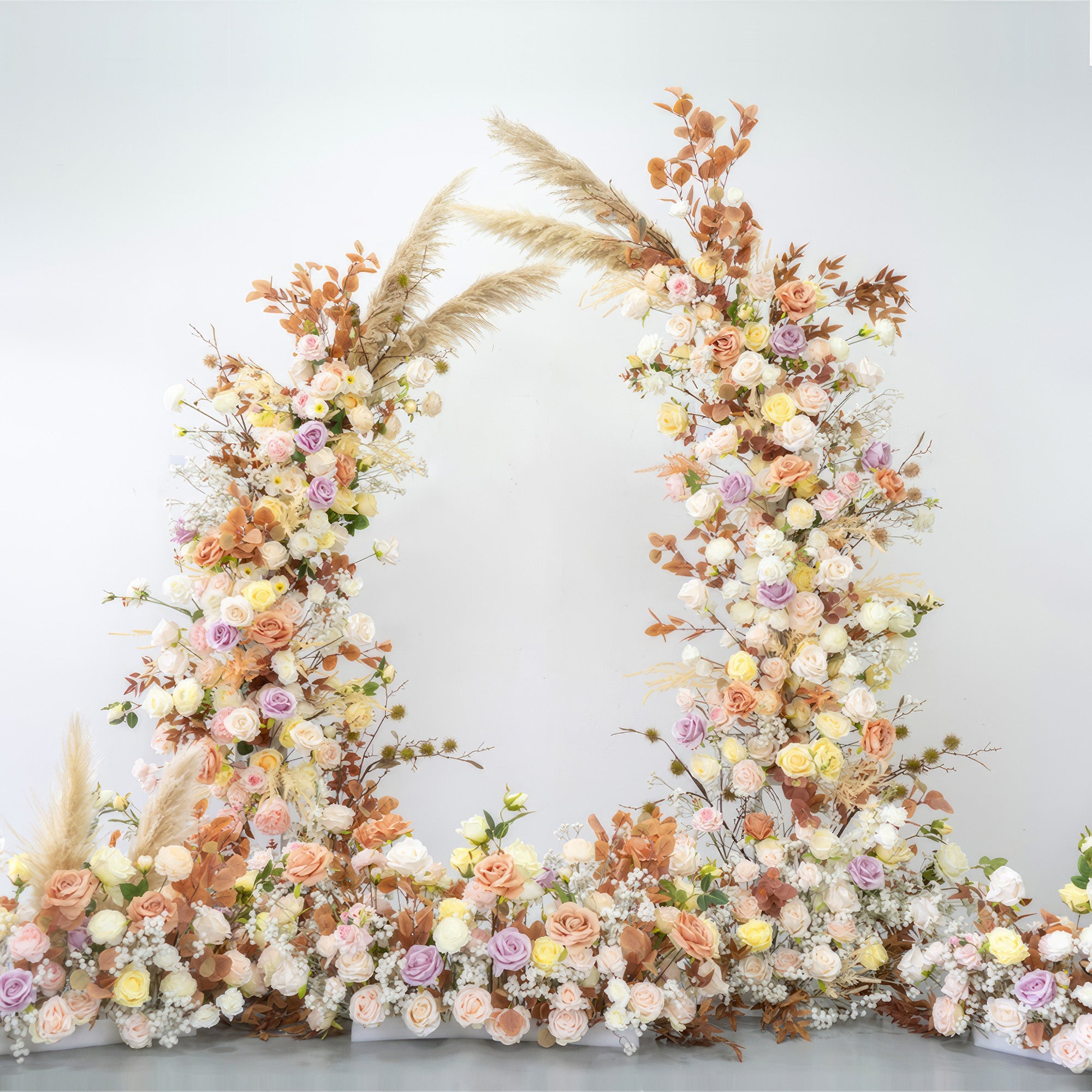 This is a floral arch composed of artificial flowers, featuring a warm and soft color palette. Shades include peach, cream, yellow, and light purple. Pampas grass and dried - looking leaves are mixed in, creating a natural, romantic, and bohemian - style decoration. 