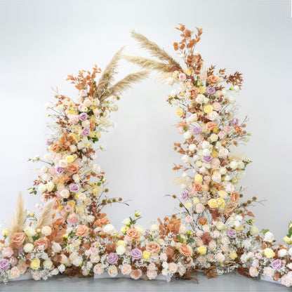 This is a floral arch composed of artificial flowers, featuring a warm and soft color palette. Shades include peach, cream, yellow, and light purple. Pampas grass and dried - looking leaves are mixed in, creating a natural, romantic, and bohemian - style decoration. 
