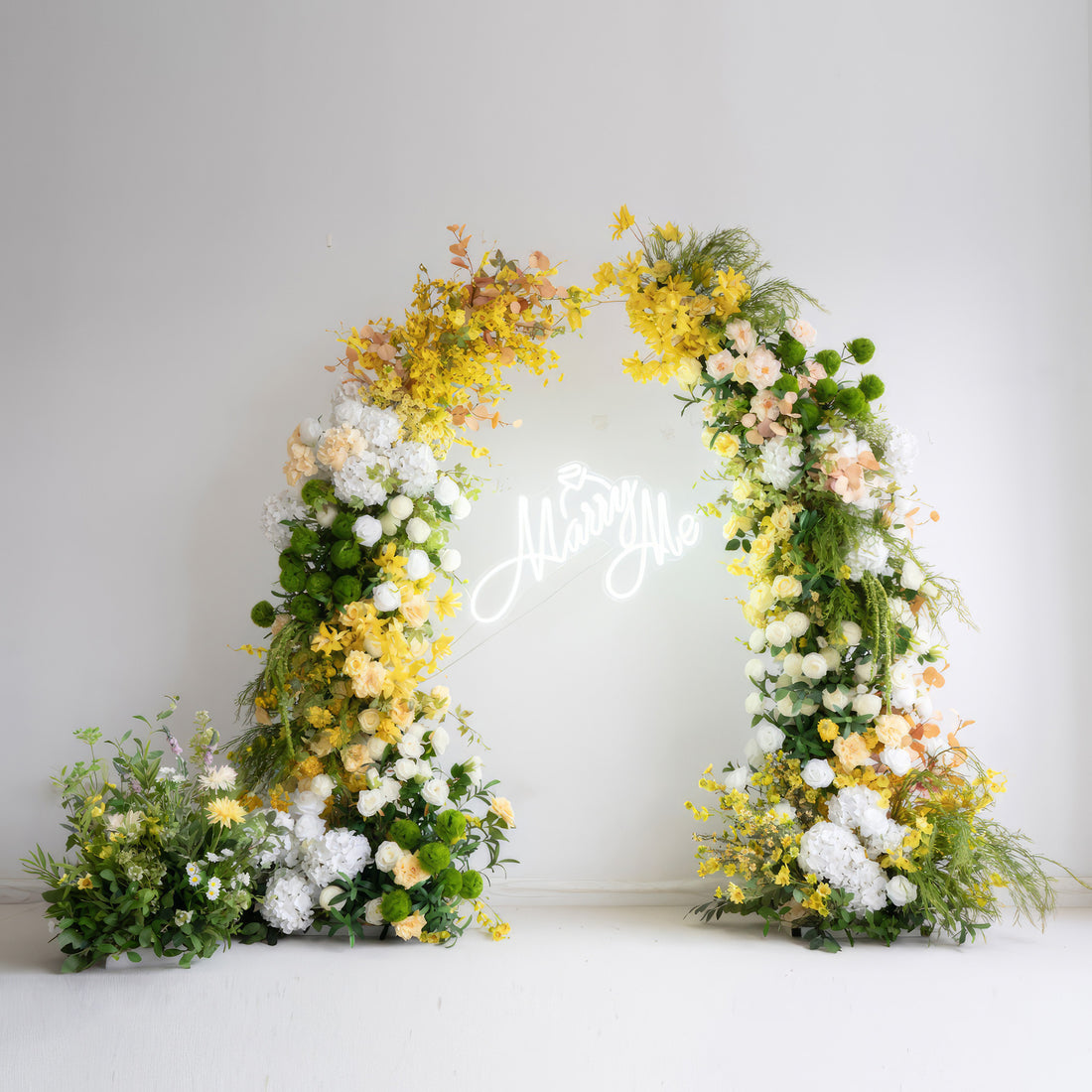 This is a floral arch made of artificial flowers, with a vivid color scheme of bright yellow, soft white, and fresh green. It includes large white hydrangeas and yellow blossoms, along with various greenery. The overall style is lively, natural, and perfect for a romantic "Marry Me" - themed setting.