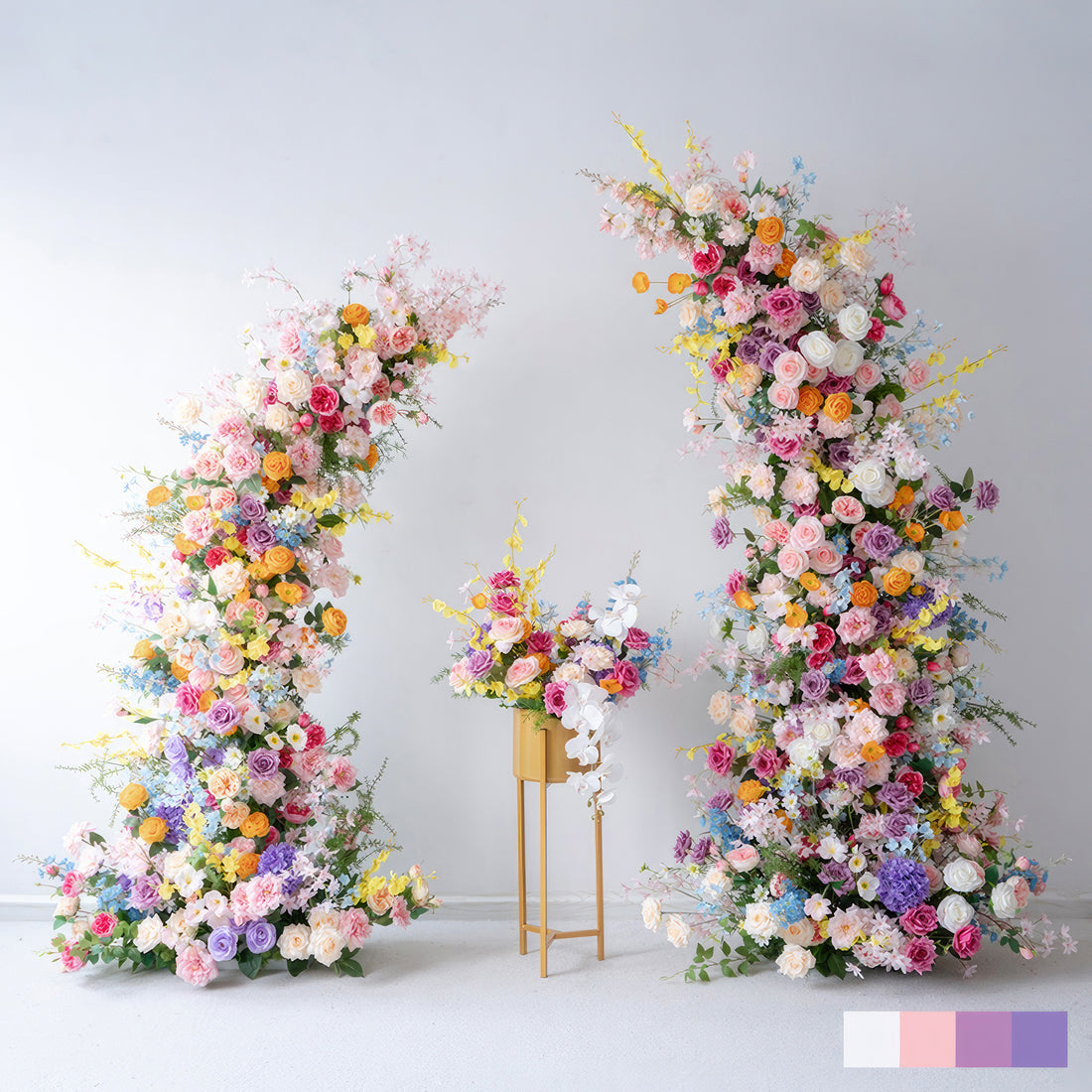 This image displays a pair of exuberant floral arches flanking a smaller floral arrangement on a stand. The arches and arrangement burst with a rainbow of colors, from soft pinks and purples to vivid oranges and yellows. They are adorned with a variety of flowers like roses, hydrangeas, and small wild - like blooms, presenting a lush, joyous, and celebratory style, ideal for festive occasions such as weddings or large parties. 