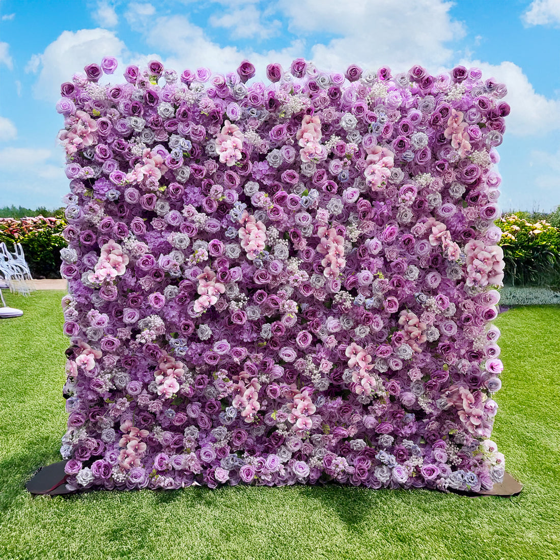 This image features a flower wall set outdoors on a grassy area. The wall is densely packed with artificial flowers, mainly roses in various shades of purple, lavender, and soft pink. There are also some smaller white flowers interspersed among the roses. The color palette creates a harmonious blend of cool tones. The style is lush and romantic, ideal for events like weddings or photo - shoots. 