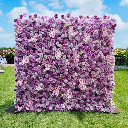 This image features a flower wall set outdoors on a grassy area. The wall is densely packed with artificial flowers, mainly roses in various shades of purple, lavender, and soft pink. There are also some smaller white flowers interspersed among the roses. The color palette creates a harmonious blend of cool tones. The style is lush and romantic, ideal for events like weddings or photo - shoots. 