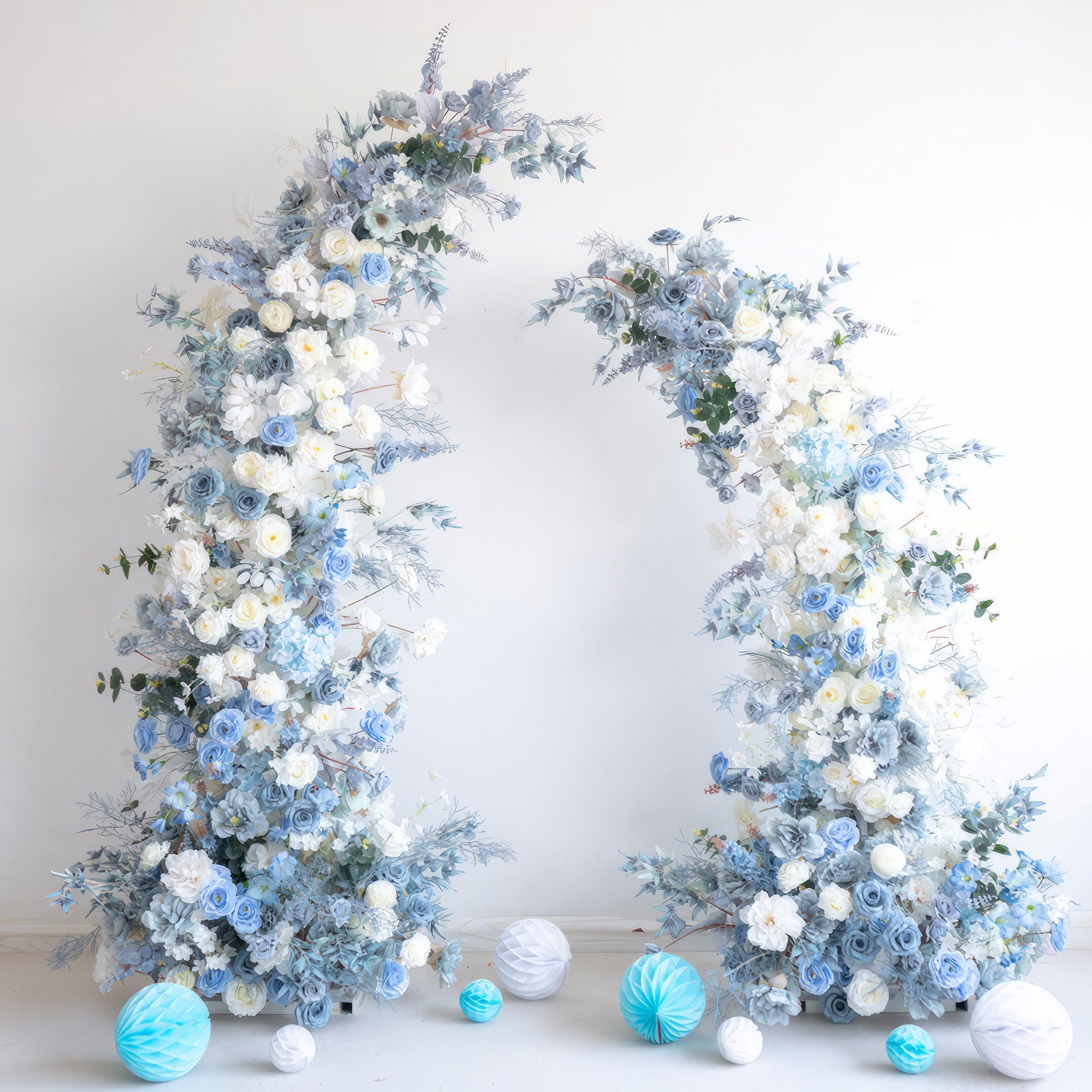 This image features a stunning floral arch made up of artificial flowers in a soothing blue - and - white color scheme. White roses and various white blooms are interspersed with different shades of blue flowers and foliage. The base is adorned with blue and white paper balls, creating an elegant, dreamy, and serene style, perfect for a wedding or a sophisticated event. 