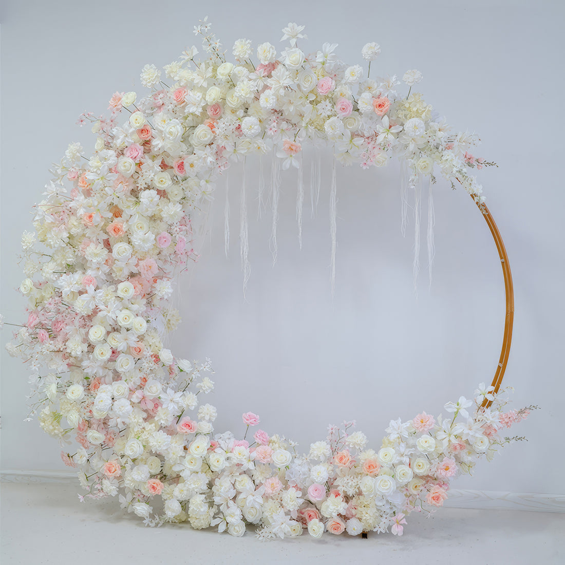 This image shows a circular floral arch adorned with artificial flowers. Colors are soft white, light pink, and peach. Flowers include roses and smaller blooms. The style is elegant and romantic, suitable for a wedding.