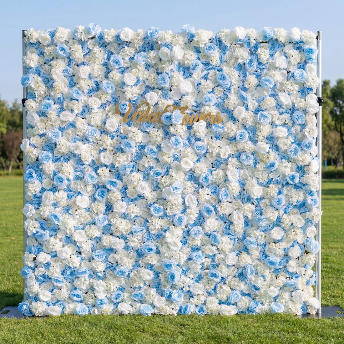 This outdoor floral backdrop (set on lush green grass, tree-framed) blends soft sky-blue and white blooms—roses and filler flowers—creating a fresh, airy display. A polished, cursive "ValarFlowers" gold sign anchors the center, contrasting vividly with the cool, serene palette. The light, elegant arrangement exudes tranquility, perfect for garden events, baby showers, or upscale photo setups with a gentle, breezy vibe.