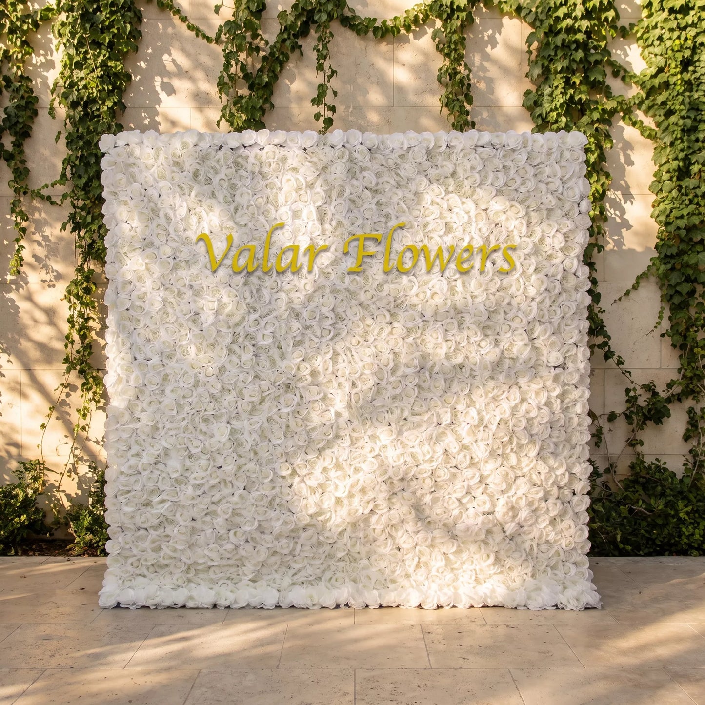 Luxury solid white artificial rose flower wall with dense full blooms, set against an ivy-covered stone courtyard wall. Glossy golden "Valar Flowers" custom branding is centered on the panel. This reusable zero-maintenance floral backdrop delivers minimalist, elegant neutral styling, ideal for wedding photo booths, baptism ceremonies, boutique brand backdrops and bright, clean social media content shoots for upscale event decor.