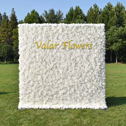 Full dense all-white artificial rose flower wall set on a sunlit green lawn with tall green trees in the background. A sleek golden custom "Valar Flowers" logo sits prominently at the center. This reusable, maintenance-free floral backdrop offers clean minimalist elegance, perfect for wedding photo booths, baptism events, boutique brand displays and neutral aesthetic social media content shoots for timeless bright decor.