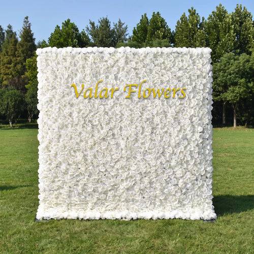 Full dense all-white artificial rose flower wall set on a sunlit green lawn with tall green trees in the background. A sleek golden custom "Valar Flowers" logo sits prominently at the center. This reusable, maintenance-free floral backdrop offers clean minimalist elegance, perfect for wedding photo booths, baptism events, boutique brand displays and neutral aesthetic social media content shoots for timeless bright decor.