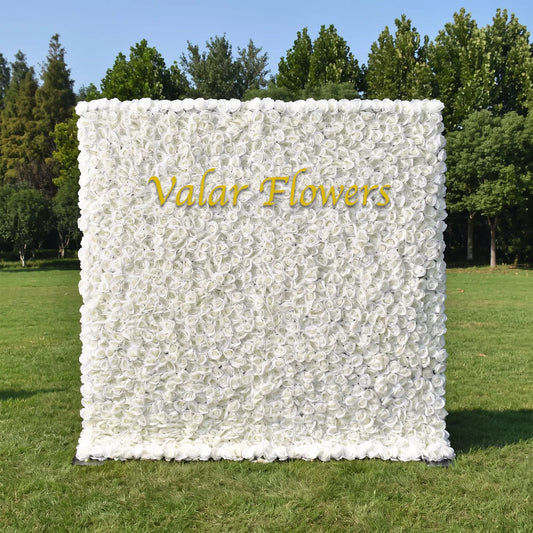 Full dense all-white artificial rose flower wall set on a sunlit green lawn with tall green trees in the background. A sleek golden custom "Valar Flowers" logo sits prominently at the center. This reusable, maintenance-free floral backdrop offers clean minimalist elegance, perfect for wedding photo booths, baptism events, boutique brand displays and neutral aesthetic social media content shoots for timeless bright decor.