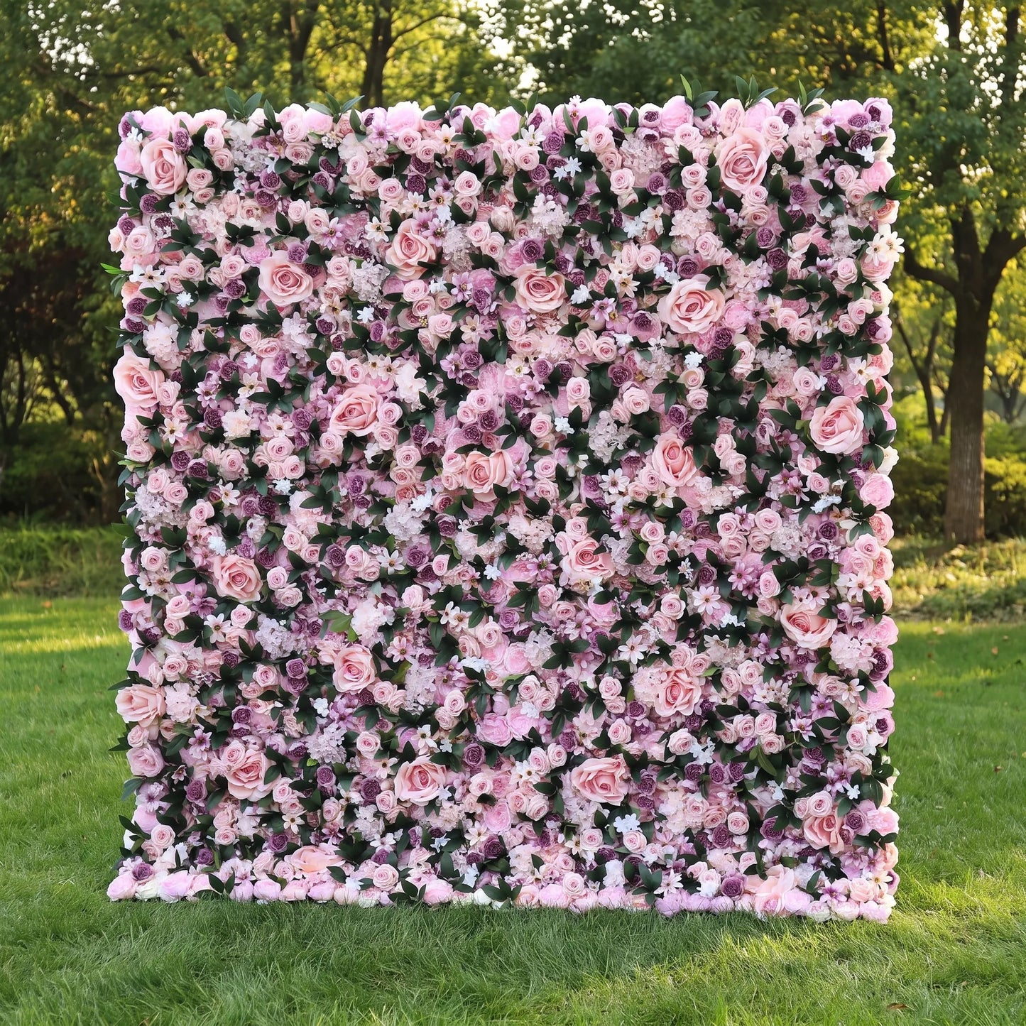 Dense luxury artificial flower wall filled with large blush roses, lavender hydrangeas, tiny purple filler blooms and fresh green foliage, displayed outdoors on a lush sunlit lawn. This reusable, maintenance-free floral panel delivers rich romantic depth, ideal for wedding photo booths, bridal parties, boutique backdrops and purple-pink themed social media shoots for dreamy, eye-catching event styling.