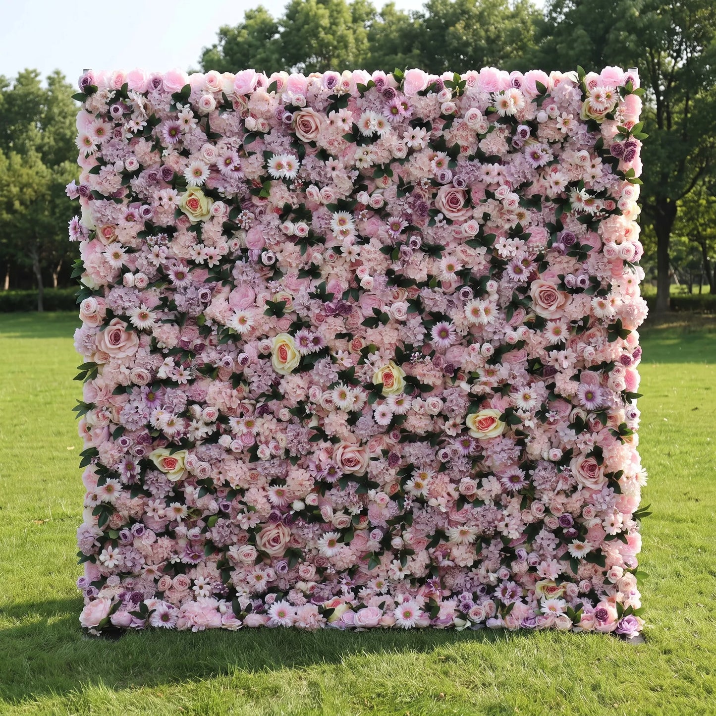 Romantic full-size artificial flower wall densely layered with blush roses, lavender hydrangeas, white daisies and soft mauve filler blooms, set on a bright green outdoor lawn. This reusable, zero-maintenance floral backdrop offers rich layered pastel texture, perfect for wedding photo booths, bridal showers, baby parties and pastel-toned social media content shoots for gentle, eye-catching celebration decor.