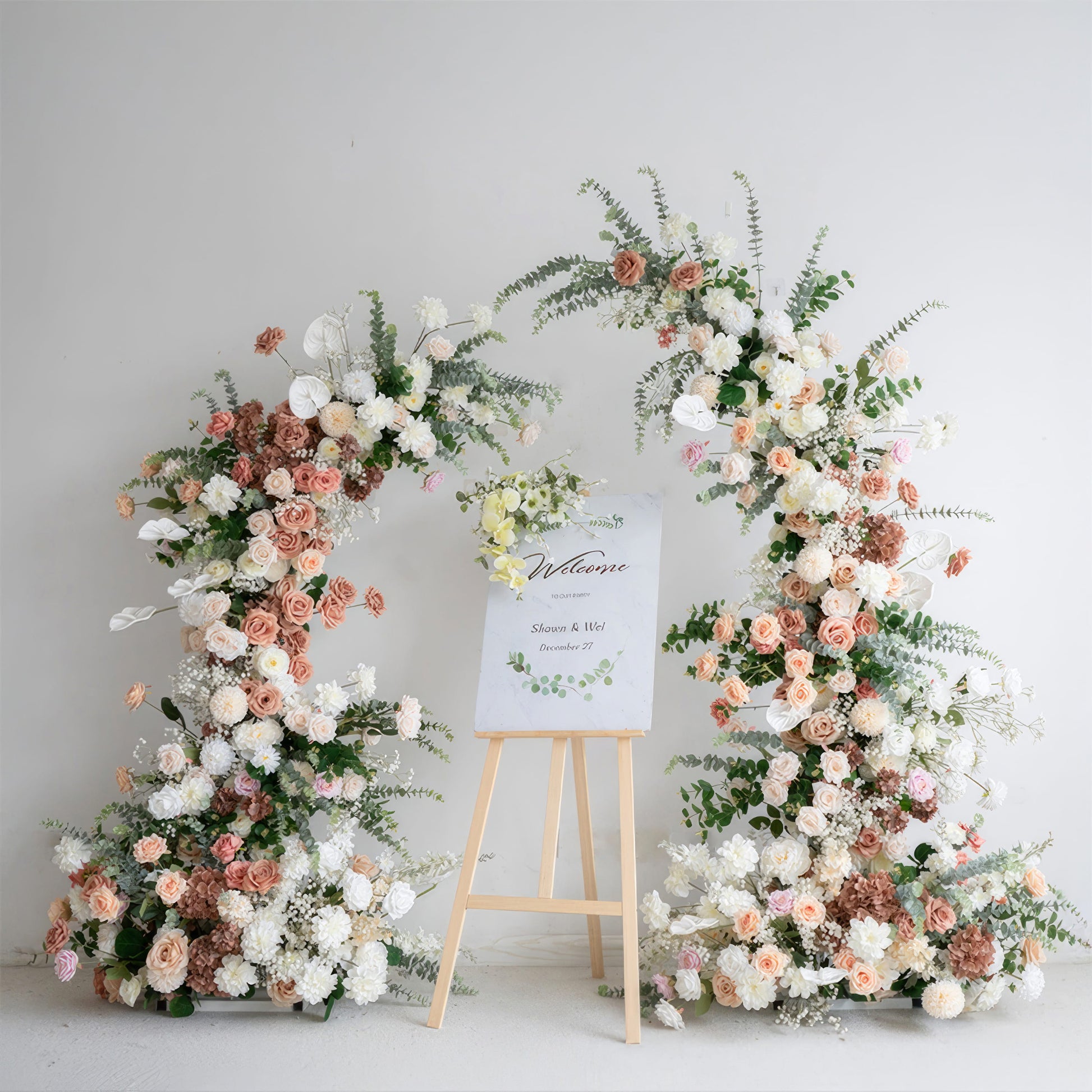A khaki color floral archway with artificial flowers, set against a white background, with a wooden sign in front displaying 'Mr & Mrs'. It's set up as a decoration for events and weddings.