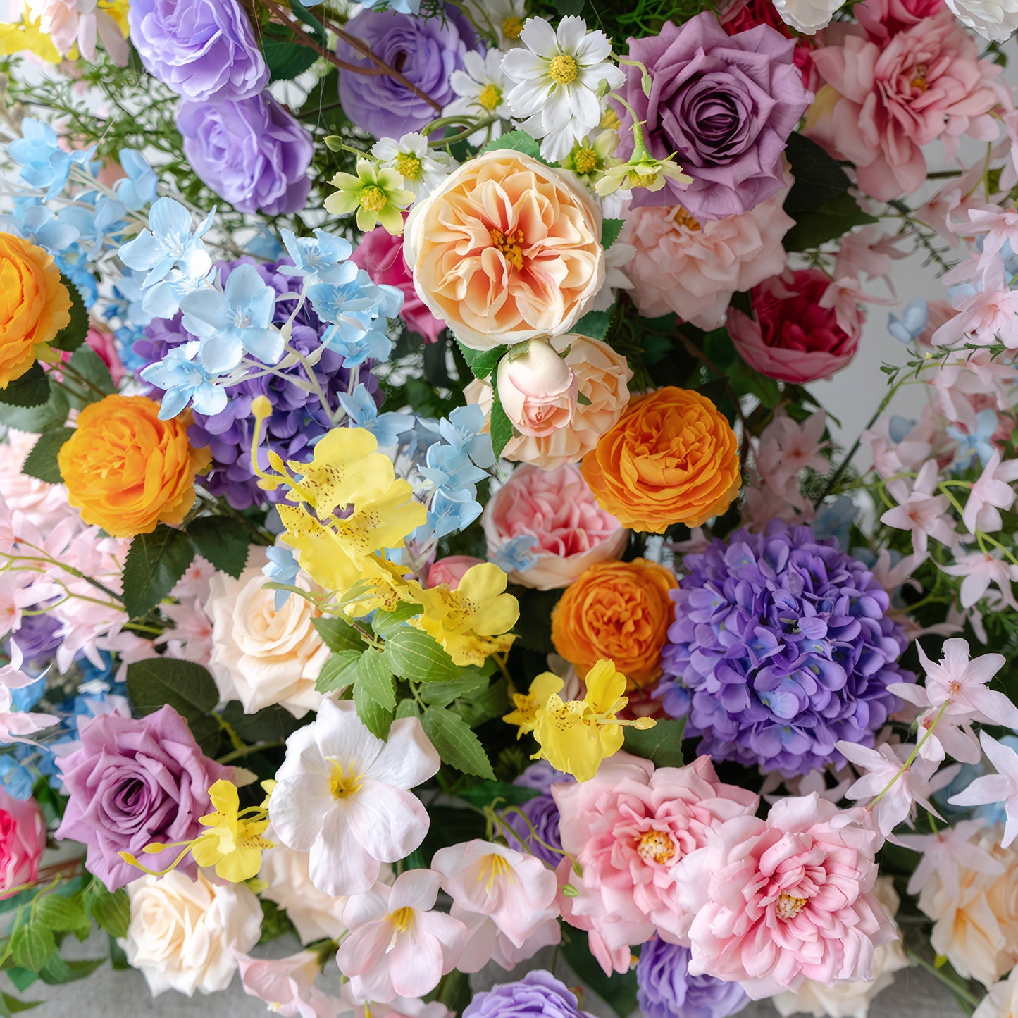 This close - up features a riot of colorful artificial flowers. There are soft pastel roses in pink, lavender, and peach, alongside vivid orange and yellow blooms. Hydrangeas in purple and blue add volume, while small white daisy - like flowers provide contrast, creating a lively, festive style. 