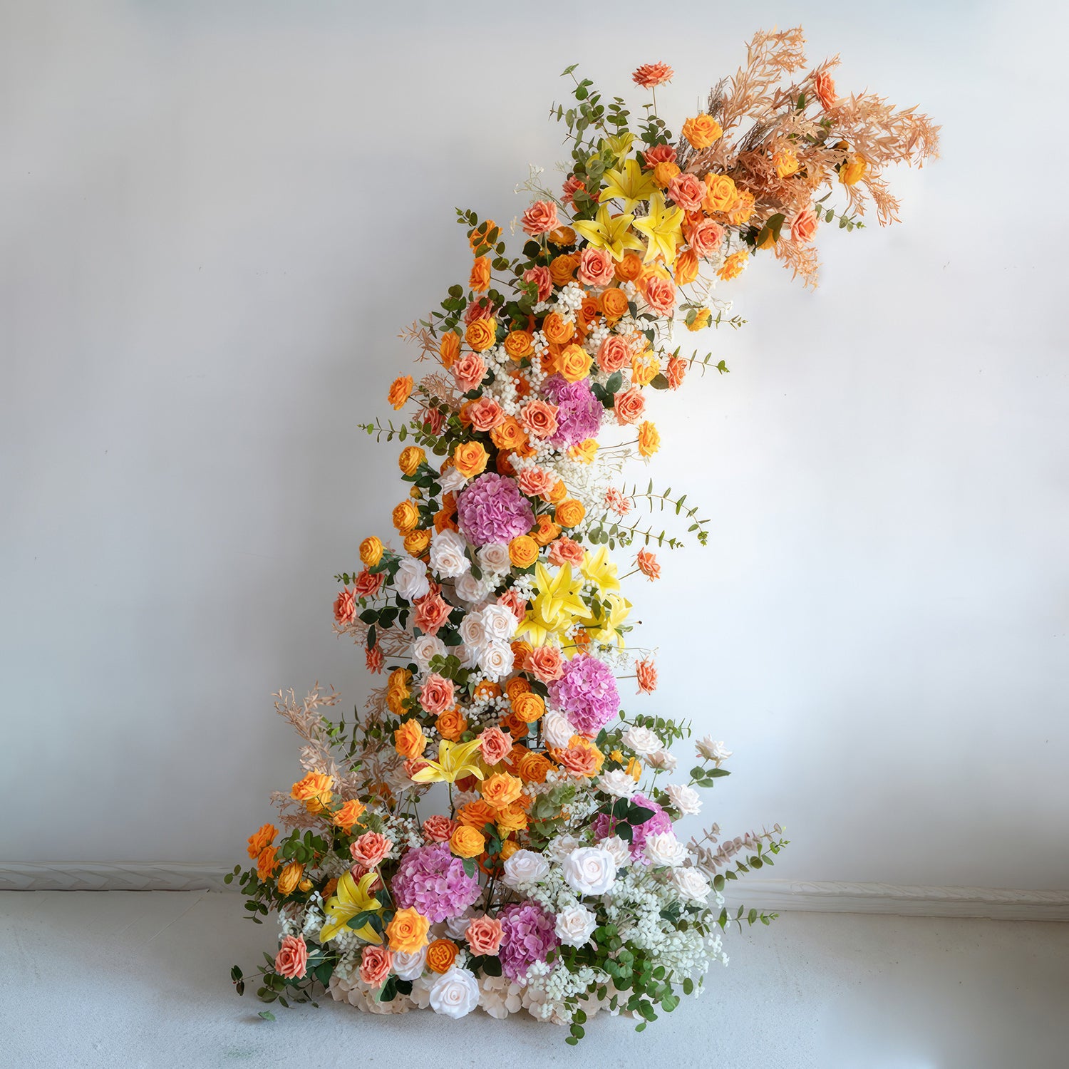 This is a stunning floral installation, likely part of an arch. It bursts with a vivid mix of colors, including bright oranges, yellows, purples, and whites. The arrangement features an assortment of flowers such as roses, hydrangeas, and lilies, along with green foliage and some dried elements. The overall effect is lively and festive, making it a great centerpiece for weddings, parties, or other celebratory events. 