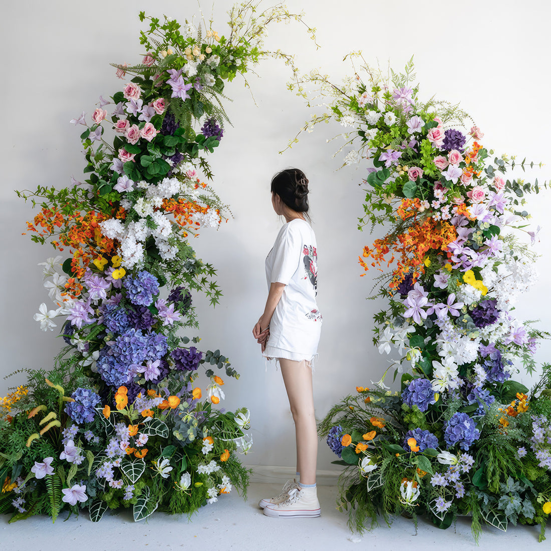 The image features a large, colorful artificial floral arch. It combines a spectrum of hues including deep purple, soft pink, bright orange, pure white, and vivid yellow. Flowers like hydrangeas and roses are visible. The style is lively and eye - catching, creating a wonderful backdrop for the person standing in front of it.