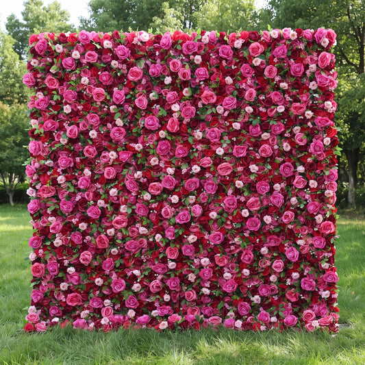 Vibrant full-panel artificial rose flower wall with rich gradient deep burgundy, fuchsia magenta and light pink roses, accented with tiny white filler blossoms and subtle green foliage, displayed on a lush sunlit lawn. This reusable, weather-resistant floral backdrop delivers bold romantic depth, ideal for wedding photo booths, anniversary parties, boutique retail displays and vivid red-pink themed social media content shoots for striking event decor.