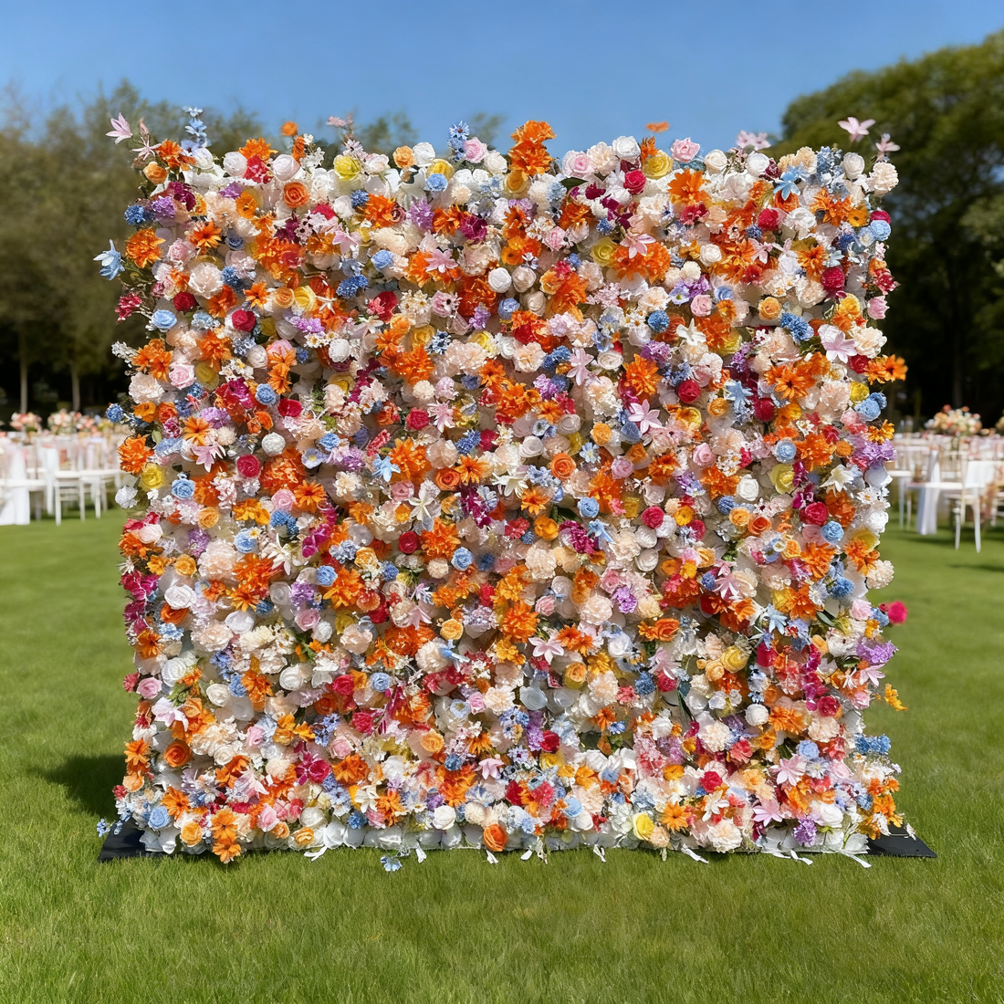 This image showcases a vibrant floral wall brimming with artificial flowers in a riot of colors, including orange, pink, blue, purple, and white. The dense arrangement features various types of blooms, creating a lively and eye - catching display. Set against an outdoor backdrop with greenery and a clear blue sky, it exudes a festive and cheerful atmosphere, perfect for events or photo backdrops. 