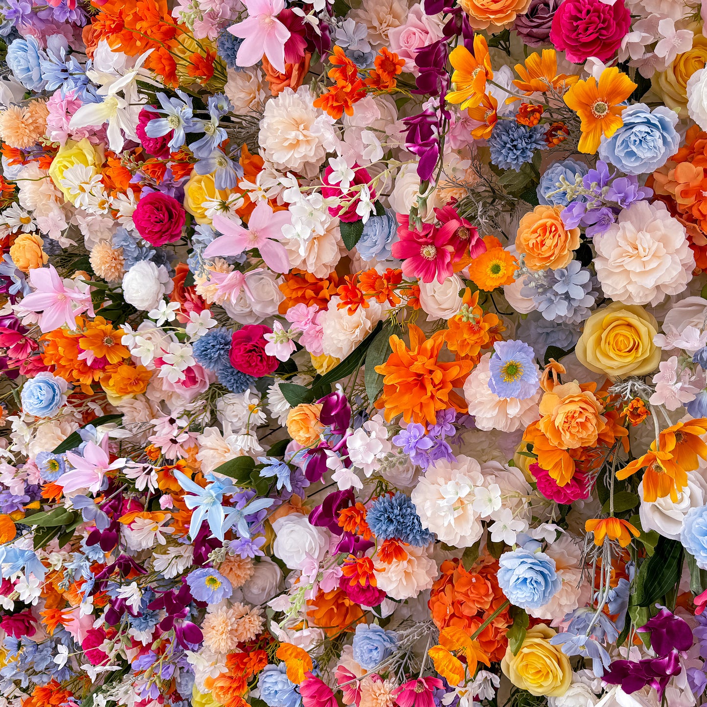 This image showcases a lush, vibrant floral arrangement densely packed with a variety of artificial flowers. The colors are a vivid mix of bright orange, soft pink, deep purple, delicate blue, creamy white, and sunny yellow. Flower types include roses, daisies, lilies - like blooms, and other assorted blossoms. The overall style is exuberant and cheerful, creating a lively and eye - catching display. 