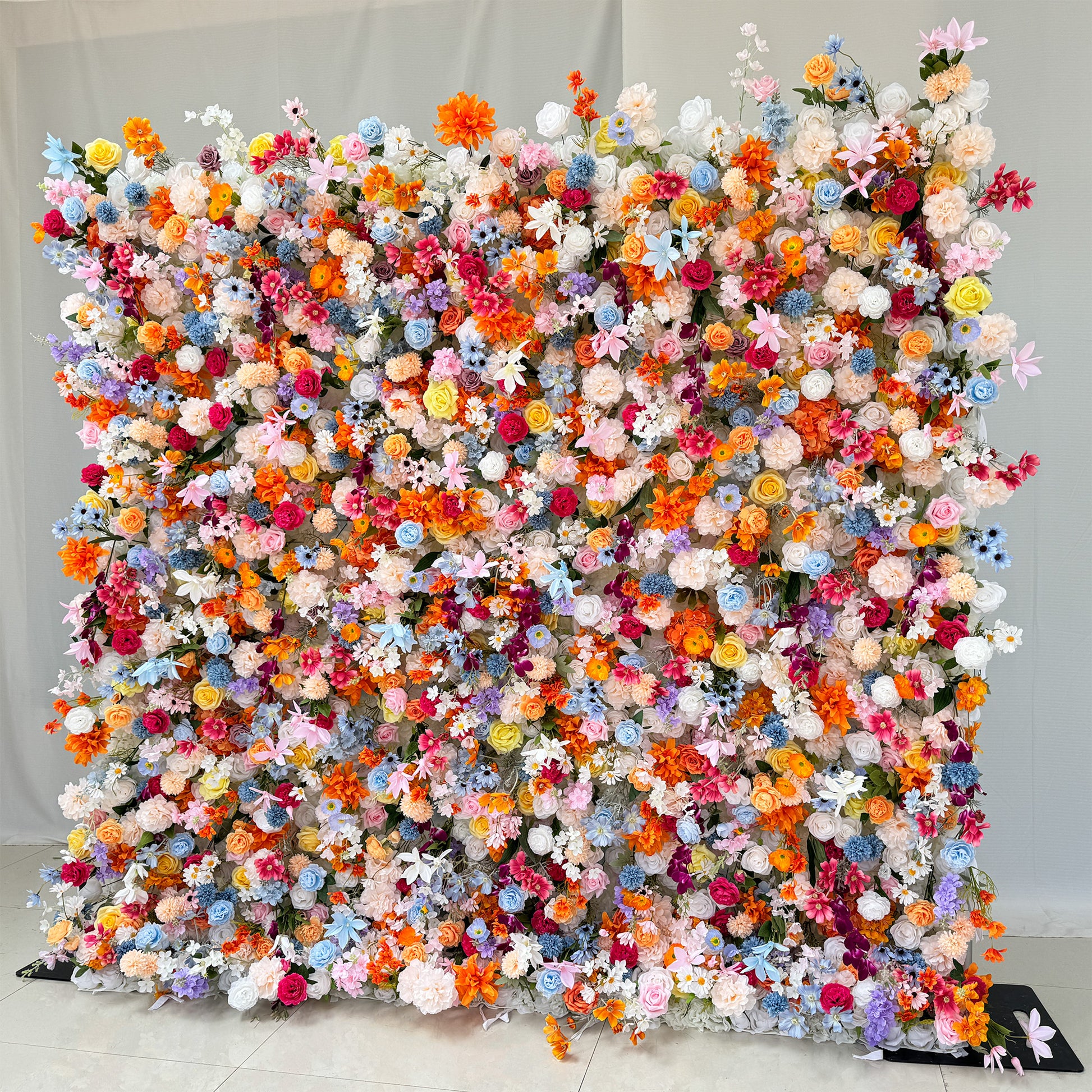 This image features a vibrant floral wall densely packed with a variety of artificial flowers. The flowers come in a vivid array of colors including bright orange, soft pink, deep purple, light blue, creamy white, and sunny yellow. Flower types include roses, daisies, and other assorted blooms, creating a lively and cheerful style. The overall arrangement is lush and eye - catching, perfect for events or photo backdrops. 