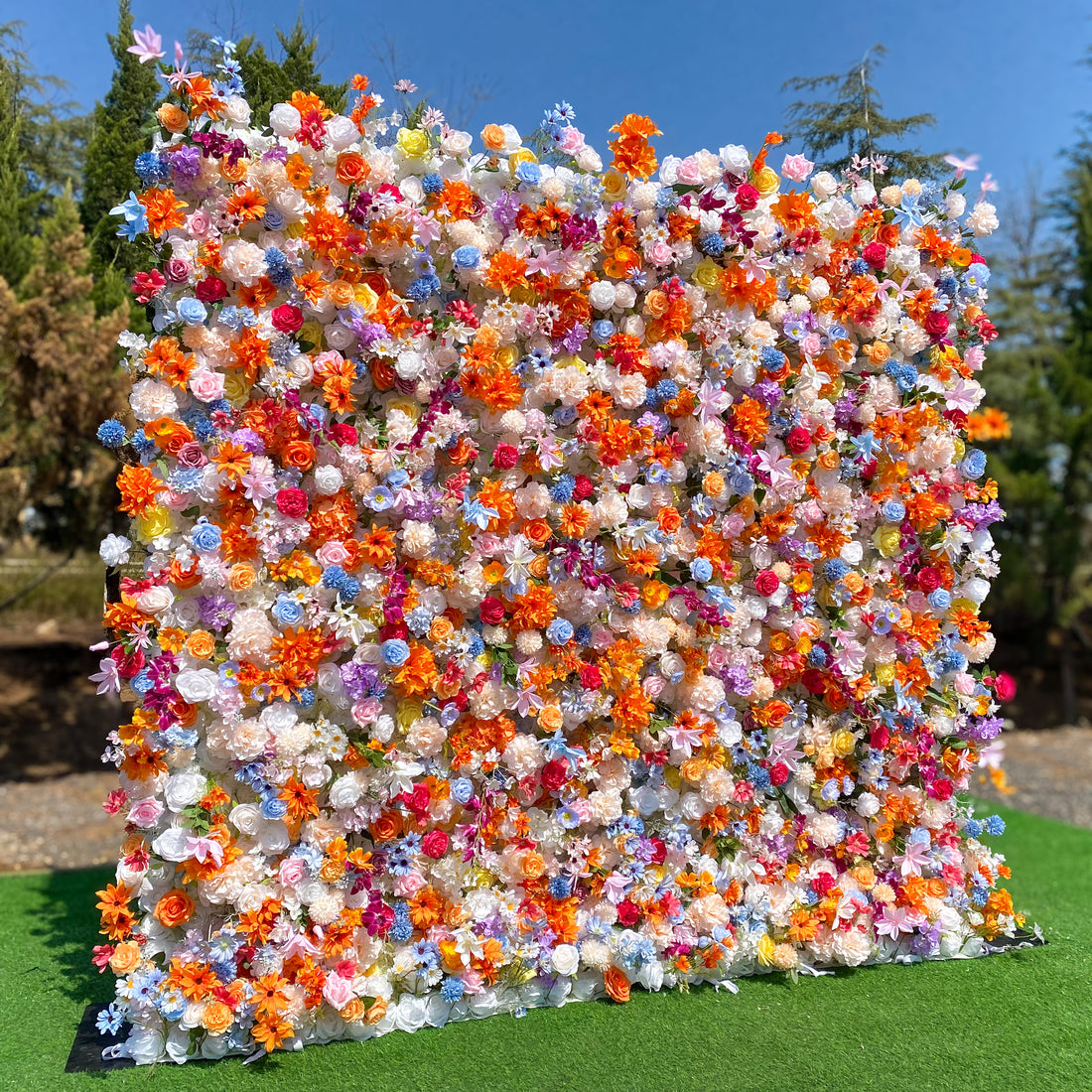 This image features a lush, vibrant floral wall densely packed with a variety of artificial flowers. The colors are a vivid mix of orange, pink, purple, blue, white, and hints of yellow. Flower types include roses, daisies, and chrysanthemum - like blooms. The overall style is exuberant and cheerful, creating a lively and eye - catching display, perfect for outdoor events or as a photo backdrop. 