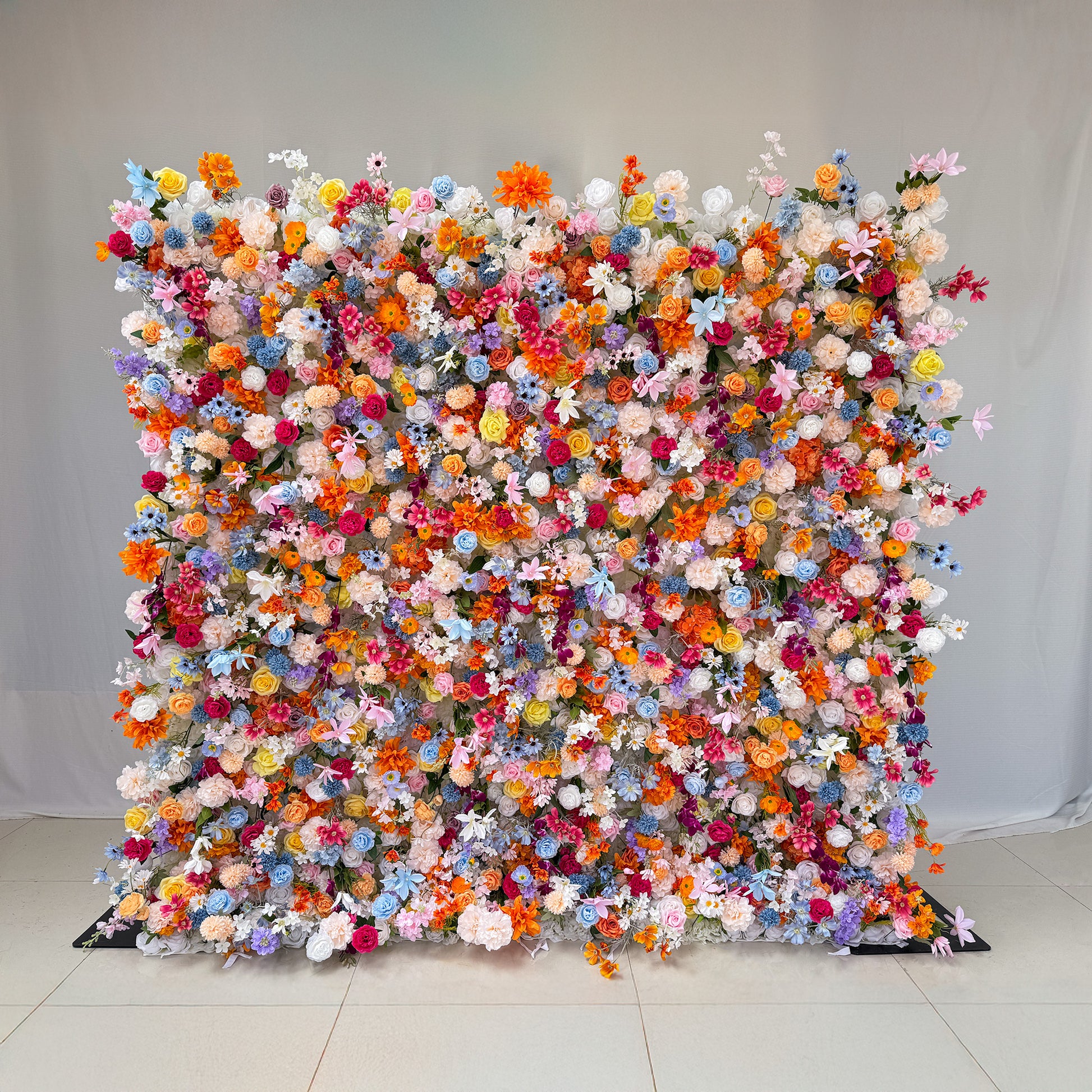 This image showcases a vibrant floral wall densely packed with artificial flowers in a kaleidoscope of colors, including orange, pink, purple, blue, white, and yellow. The flowers appear to be a mix of roses, daisies, and other assorted blooms. The overall style is lively and exuberant, creating a cheerful and eye - catching display, perfect for events or photo backdrops. 