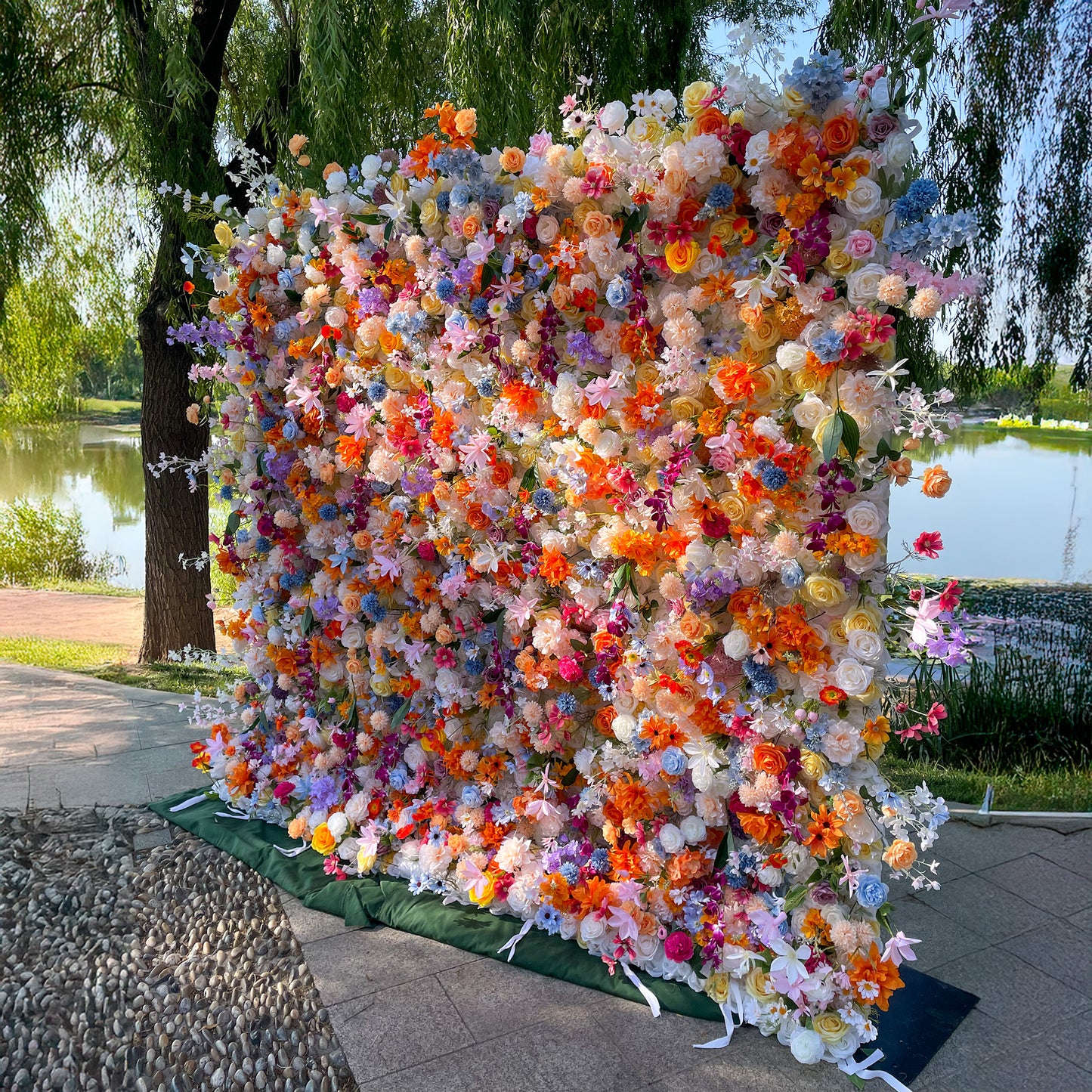 This image showcases a vibrant, lush floral wall set against a serene lakeside backdrop with willow trees. The wall is densely adorned with an array of artificial flowers in a riot of colors, including vivid oranges, soft pinks, purples, blues, and whites. Flowers like roses, daisies, and hydrangea - lookalikes are interspersed, creating a lively, cheerful, and whimsical style, perfect for photo - taking. 