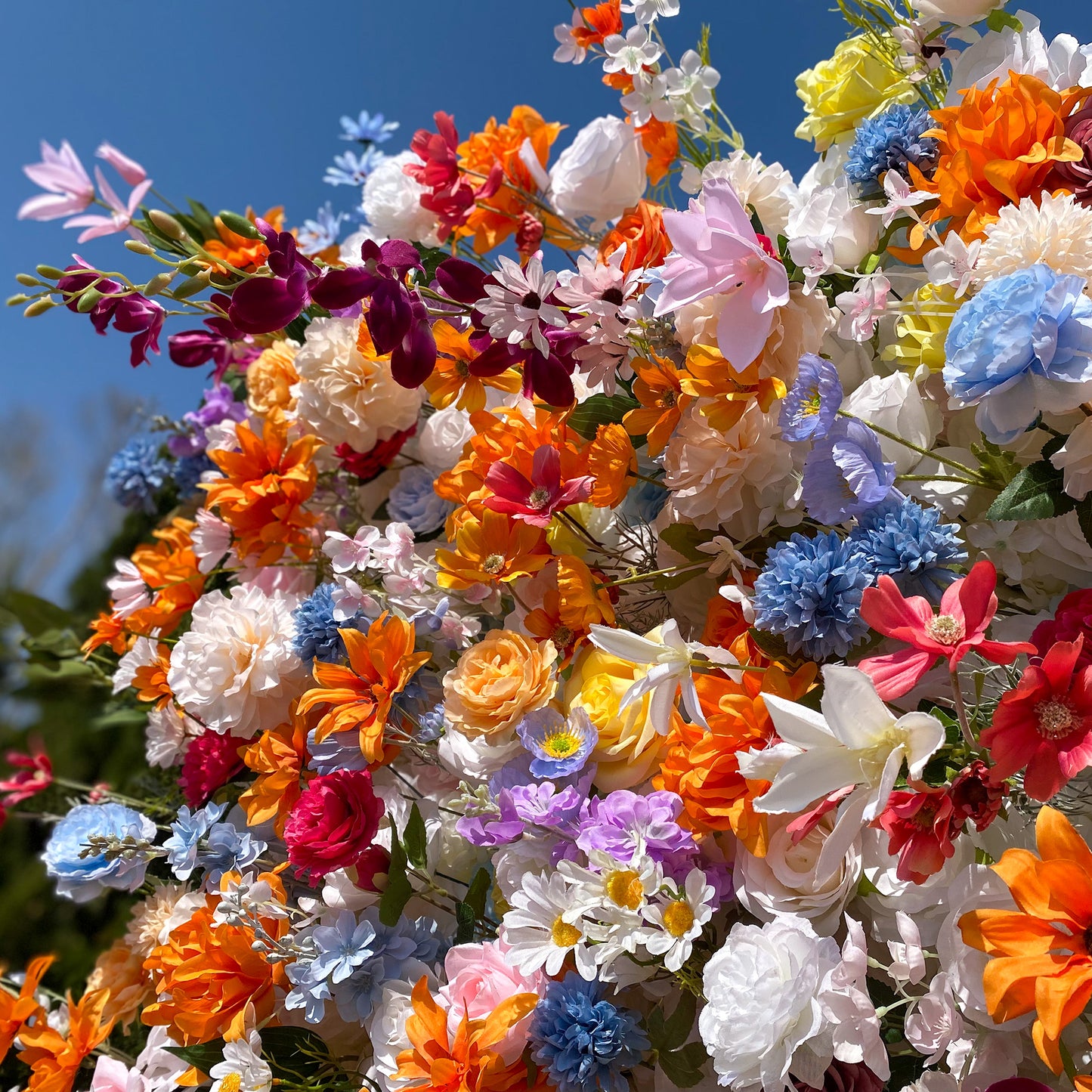 This image showcases a dazzling assortment of artificial flowers set against a clear blue sky. The flowers display a vivid spectrum of colors, including bright orange, deep red, soft pink, gentle lavender, and pure white. Varieties such as roses, daisies, and lilies - like blooms are interwoven, creating a lively and whimsical style that exudes a sense of joy and celebration. 
