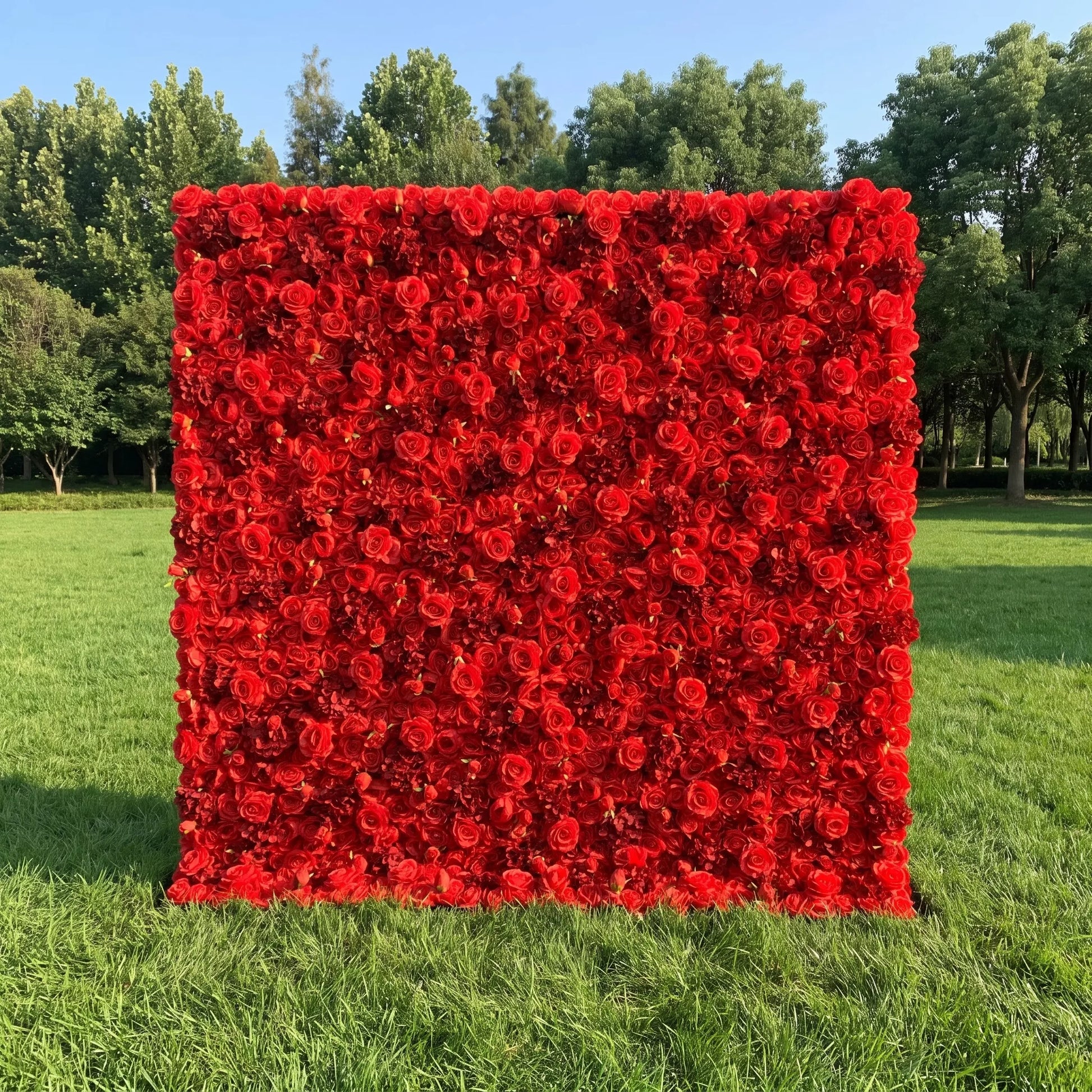 Stunning solid bright red artificial rose flower wall with dense full bloom coverage, placed on a sunlit green lawn backed by lush tall trees. This reusable, weatherproof floral backdrop delivers bold romantic drama, perfect for wedding photo booths, Valentine’s events, anniversary parties, boutique retail displays and vivid red-themed social media content shoots for eye-catching statement event decor.