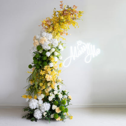 This is an artificial flower arrangement forming a curved structure, boasting a vibrant color palette of bright yellow, pure white, and rich green. It includes large white hydrangeas, yellow flowers, and spherical greenery. With the &quot;Marry Me&quot; neon sign in the background, it conveys a romantic and fresh style. 