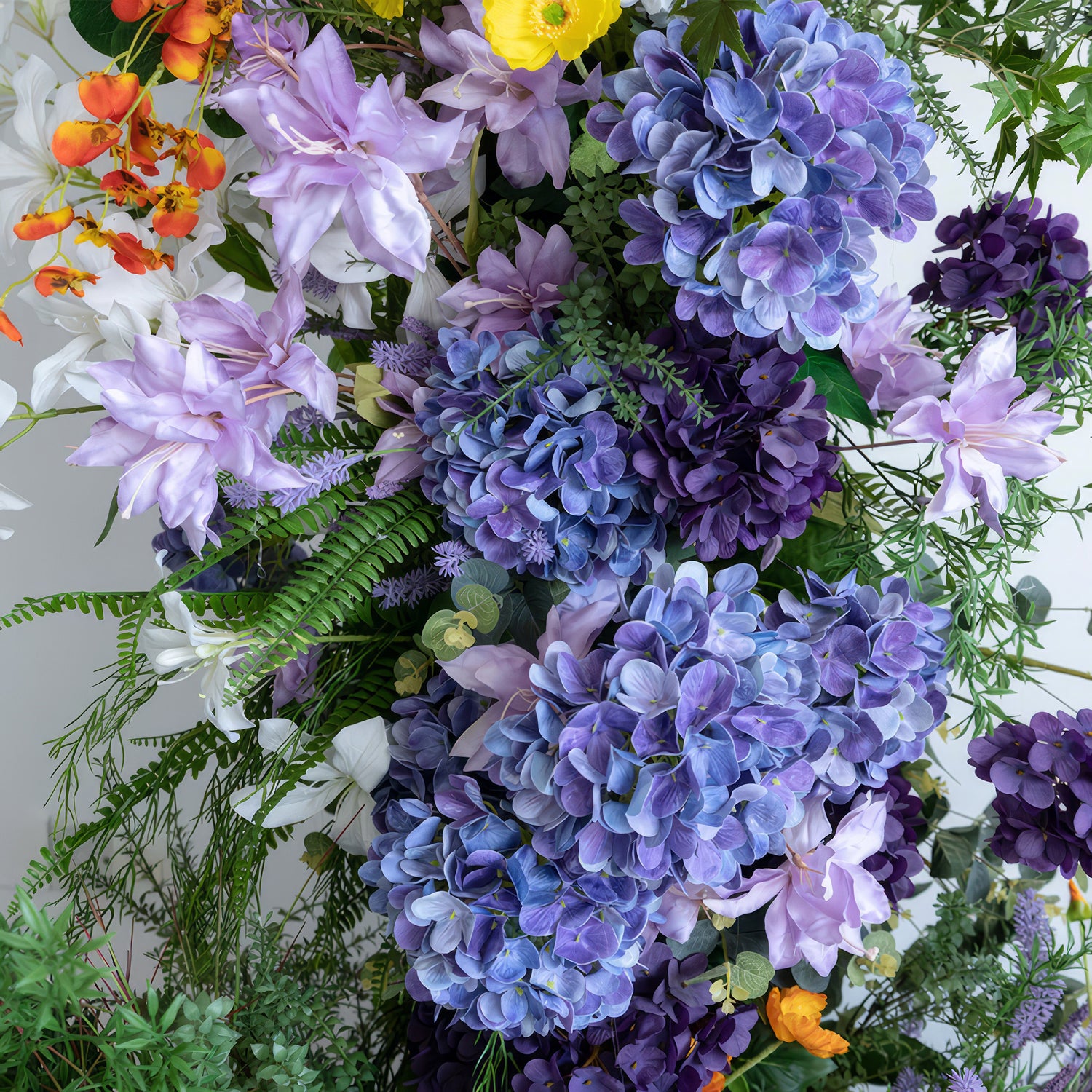 This is a close - up of an artificial flower arrangement. The color palette includes shades of purple from light lavender to deep violet, along with white, orange, and yellow. It features large hydrangeas and delicate, broad - petaled purple flowers, creating a rich and vibrant style. 