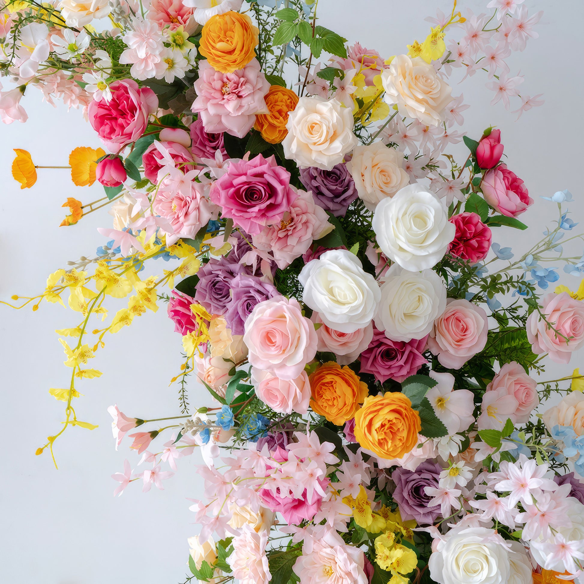 This floral arrangement is a vibrant medley of colors. It includes roses in shades like white, pink, magenta, lavender, and orange. Smaller flowers in yellow and light blue add variety. With green foliage, it has a lively, celebratory style, perfect for joyous events. 