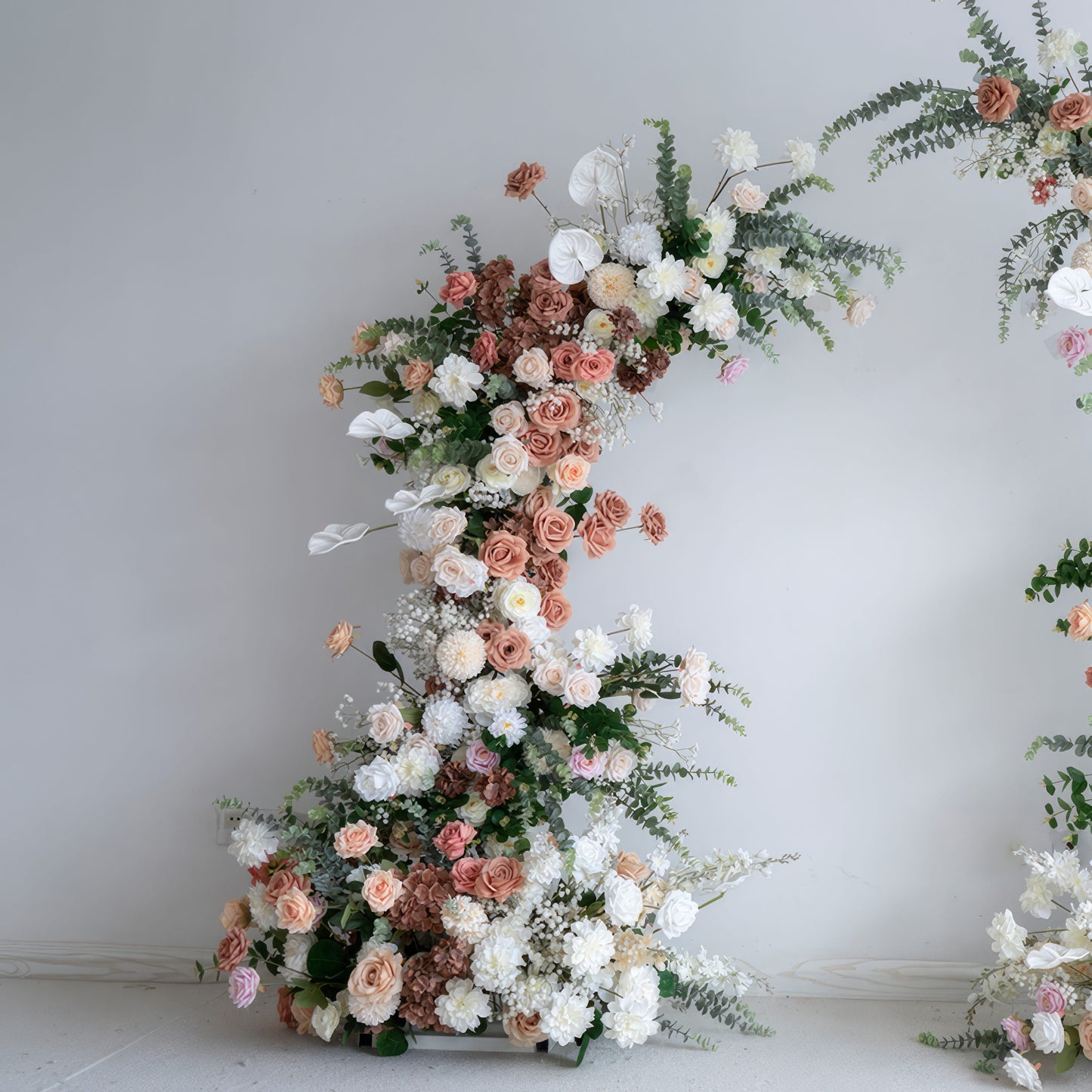 A romantic, asymmetrical floral arch from **Valar Flowers**, crafted with a soft, warm palette of blush pink, terracotta, and creamy white blooms—including roses, peonies, and delicate baby's breath—intertwined with silvery eucalyptus and lush greenery. Set against a neutral studio backdrop, it creates a dreamy, elegant, and sophisticated aesthetic ideal for luxury weddings and editorial photoshoots.
