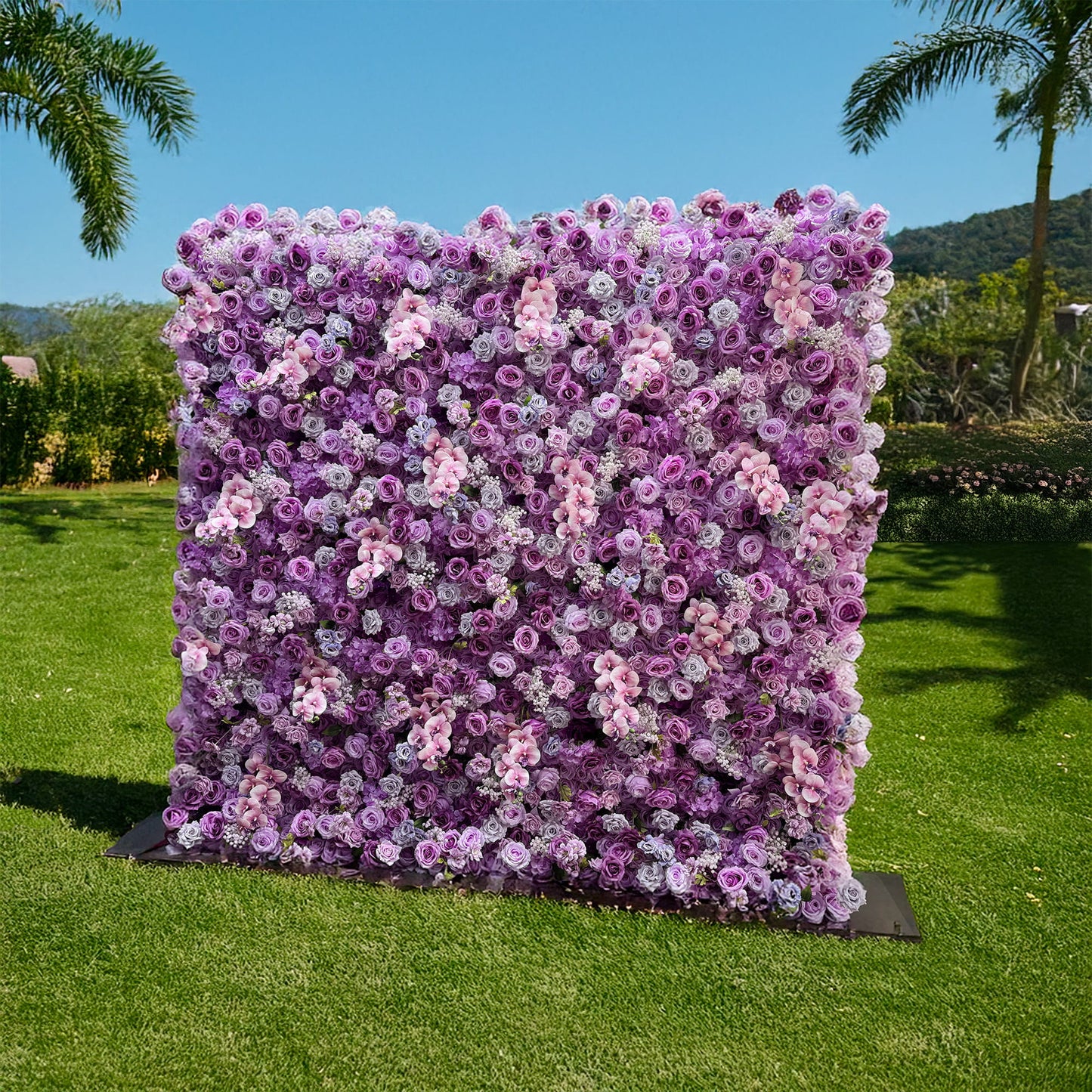 This image depicts a flower wall set against a lush green lawn, with palm trees and a clear blue sky in the background. The wall is densely covered with artificial flowers, mainly roses in various shades of purple, lavender, and soft pink. Smaller clusters of white flowers are interspersed among the roses. The overall style is lush and romantic, perfect for events like weddings or photo - shoots. 
