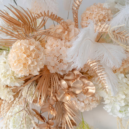 This image showcases an artificial flower arrangement with hydrangeas, palm fronds, and orchids. In soft white, peach, and metallic gold tones, it has a luxurious and contemporary style. 