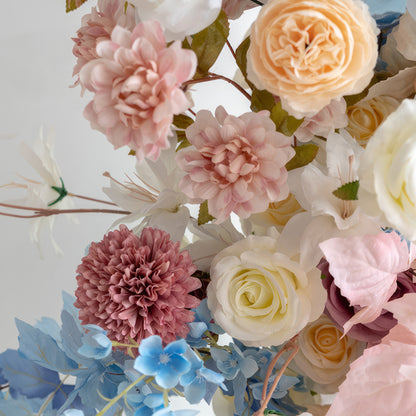 This floral arrangement features a charming mix of colors including soft pinks, creamy whites, warm peaches, and cool blues. With roses, dahlias and other blooms, it offers a sweet, delicate style, perfect for adding a touch of beauty to events like weddings or home decor. 