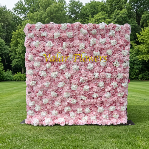 Lush blush pink artificial flower wall filled with soft pink roses and scattered white hydrangea blooms, displayed on a smooth green lawn with leafy trees behind. Glossy golden "Valar Flowers" custom branding sits front and center. This reusable, maintenance-free floral backdrop offers gentle romantic texture, perfect for wedding photo booths, bridal showers, baby girl parties and pink-toned social media content shoots for soft, elegant event styling.