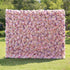 This outdoor floral backdrop rests on a sunlit green lawn, framed by leafy, sun-dappled trees. It is a dense, textured wall of artificial blooms in soft blush pink, ivory, and subtle peach tones, featuring tightly packed roses and delicate filler flowers that create a dreamy, romantic surface. The gentle gradient palette and uniform, long-lasting artificial construction make it ideal for garden weddings, proposal setups, or photo booths seeking a feminine, timeless focal point.